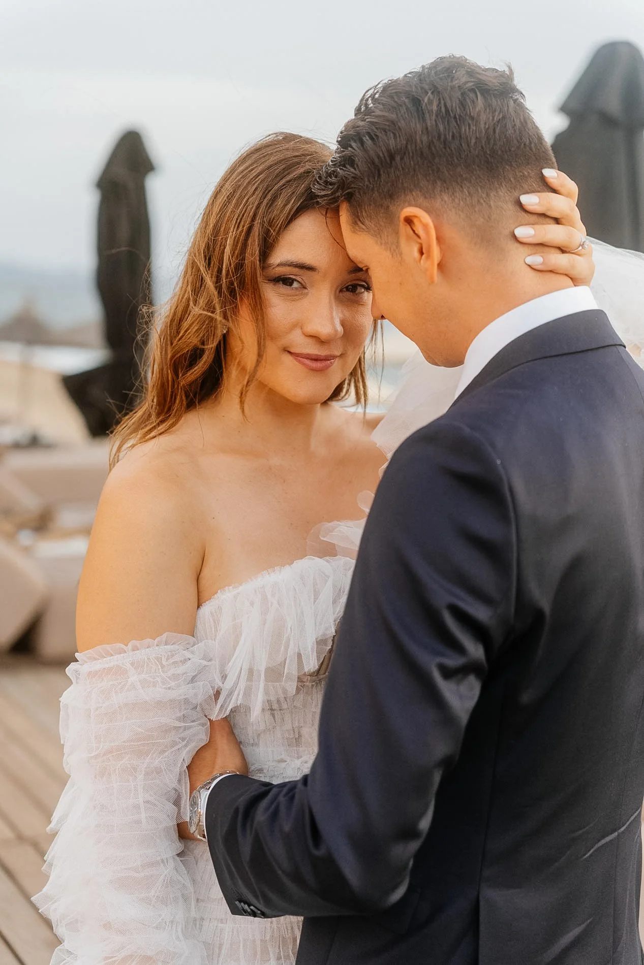 A bride and groom sharing a close moment during their wedding, with the bride smiling softly and the groom's forehead touching hers, outdoors with umbrellas in the background.