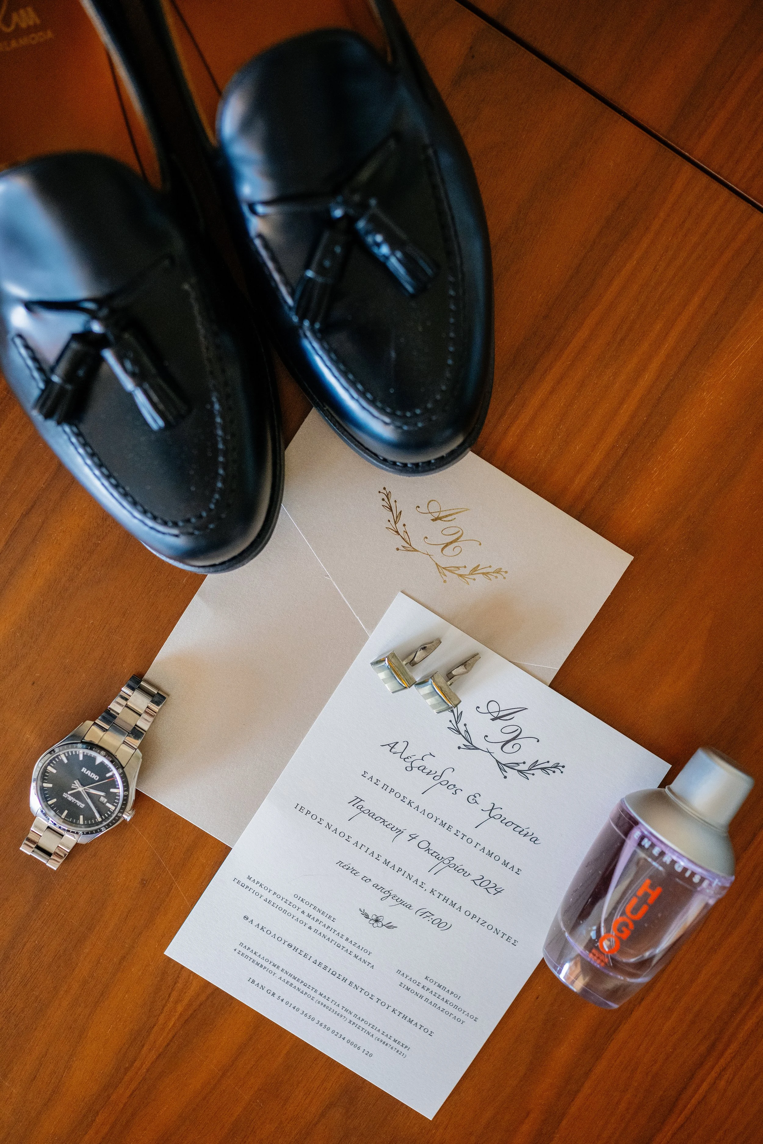 Black formal shoes, a silver wristwatch, wedding invitation cards with gold designs, a perfume bottle, and a hair spray bottle arranged on a wooden surface.