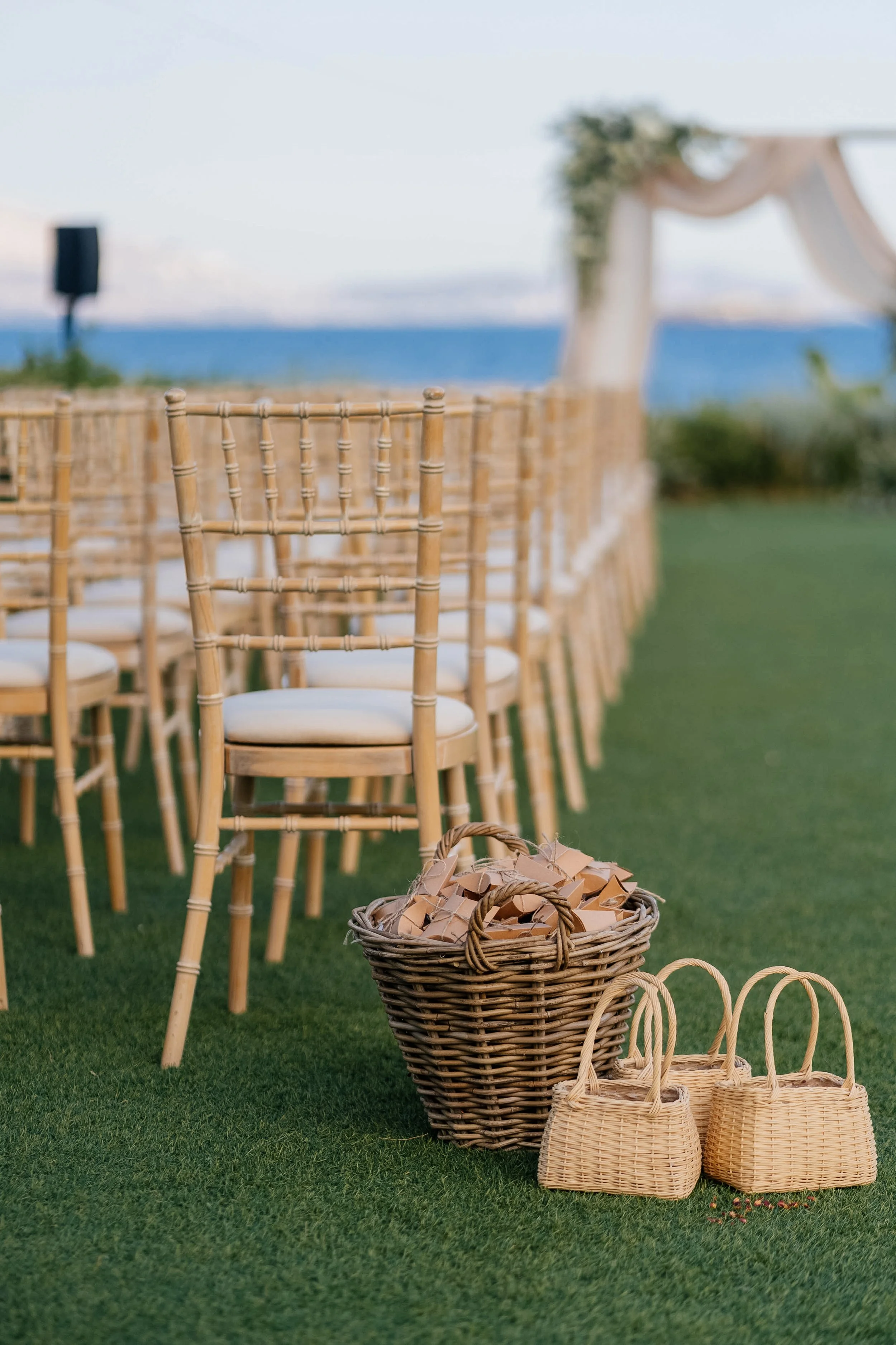Bamboo chairs arranged in rows on a green lawn, with several wicker baskets filled with small paper tags or cards in front, outdoors near a body of water with a blurred background of an archway decorated with fabric and greenery.