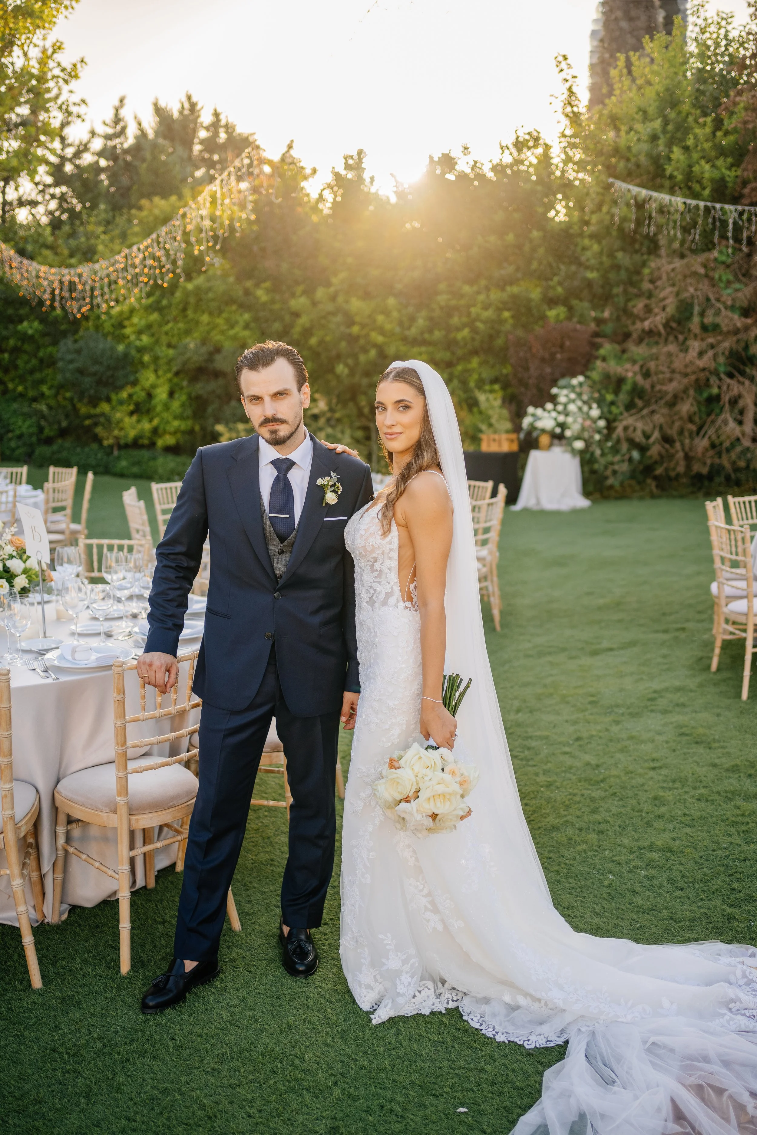 A bride and groom standing together outdoors during a wedding reception in the evening. The groom is wearing a navy blue suit and tie, and the bride is dressed in a white lace wedding gown and holding a bouquet of white and peach roses. There are dec