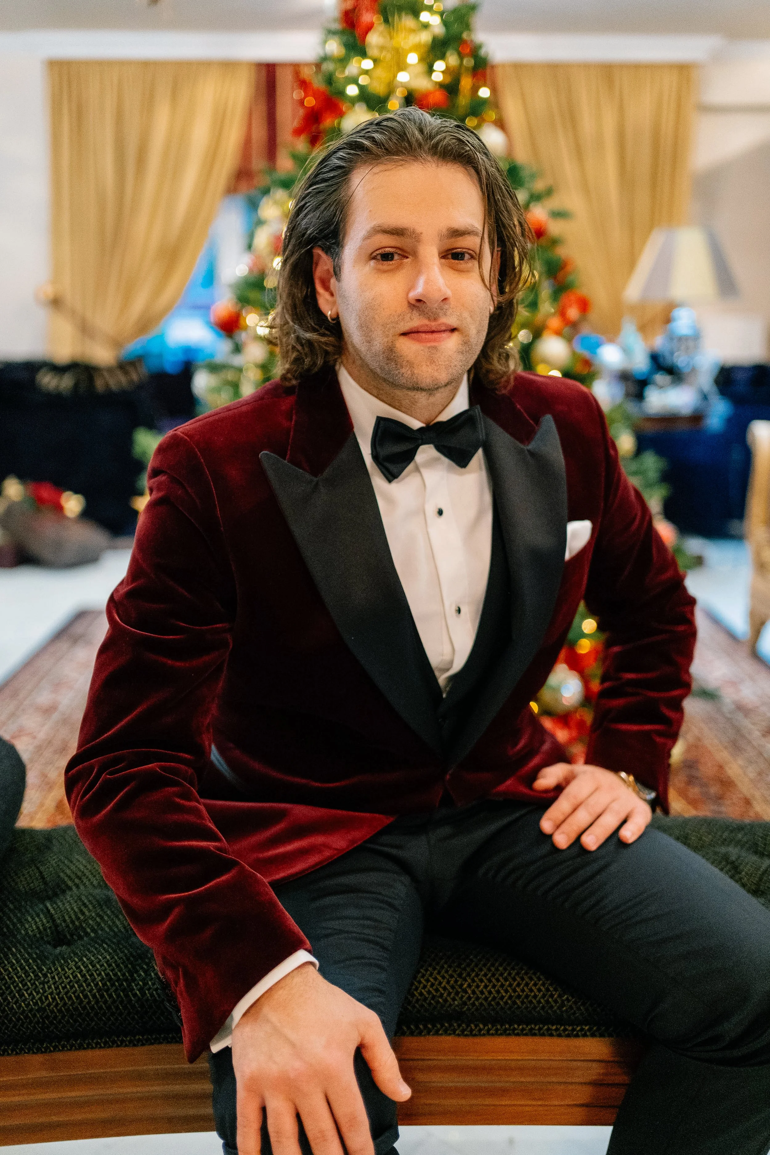 A man with long, dark hair dressed in a tuxedo with a red velvet blazer and black bow tie, sitting in front of a decorated Christmas tree.