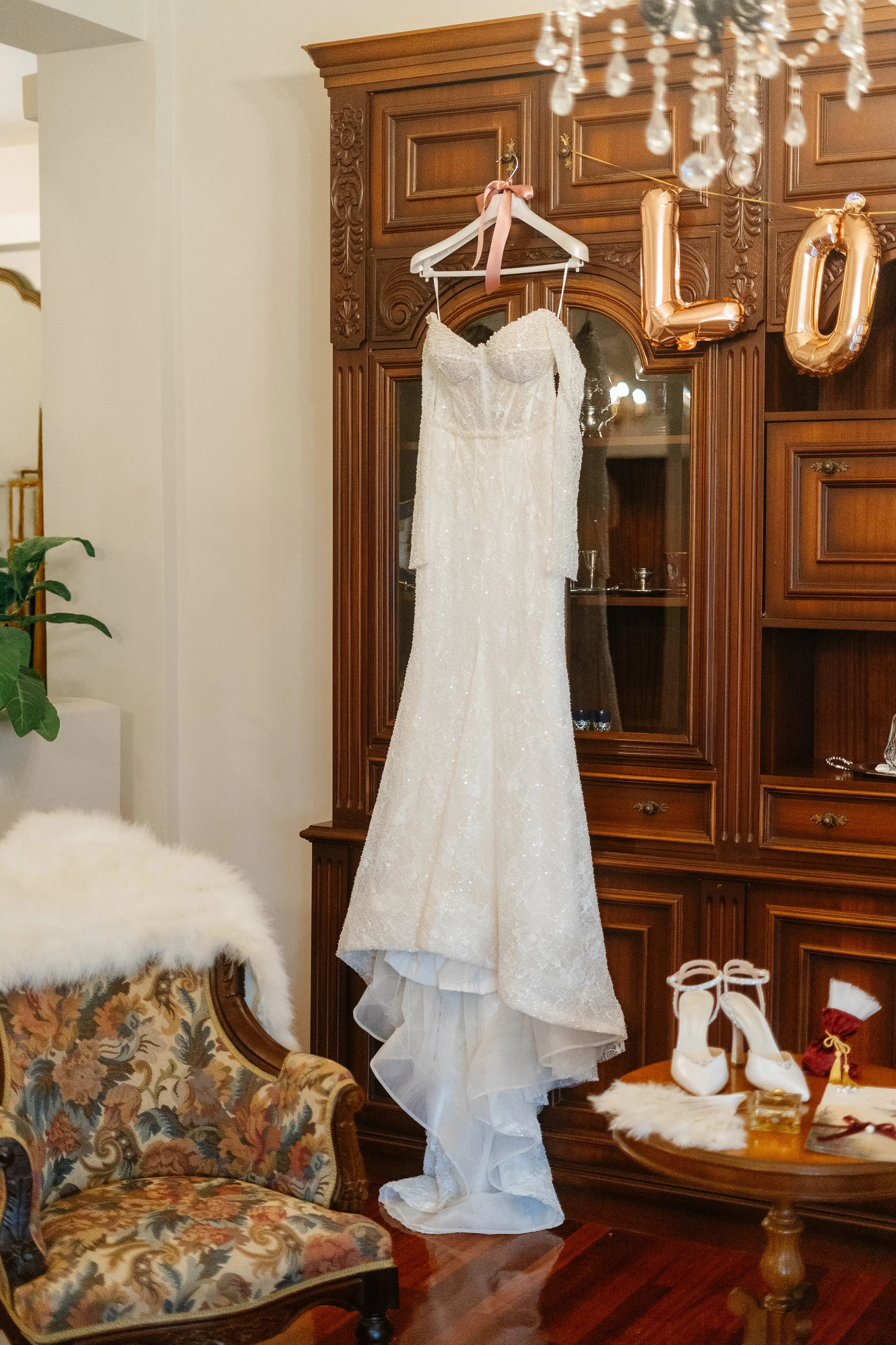 A wedding dress hanging on a hanger with a pink ribbon, displayed in a room with a wooden cabinet, a chandelier, a floral upholstered chair, white bridal shoes, a small red gift bag, and a gold box on a table.