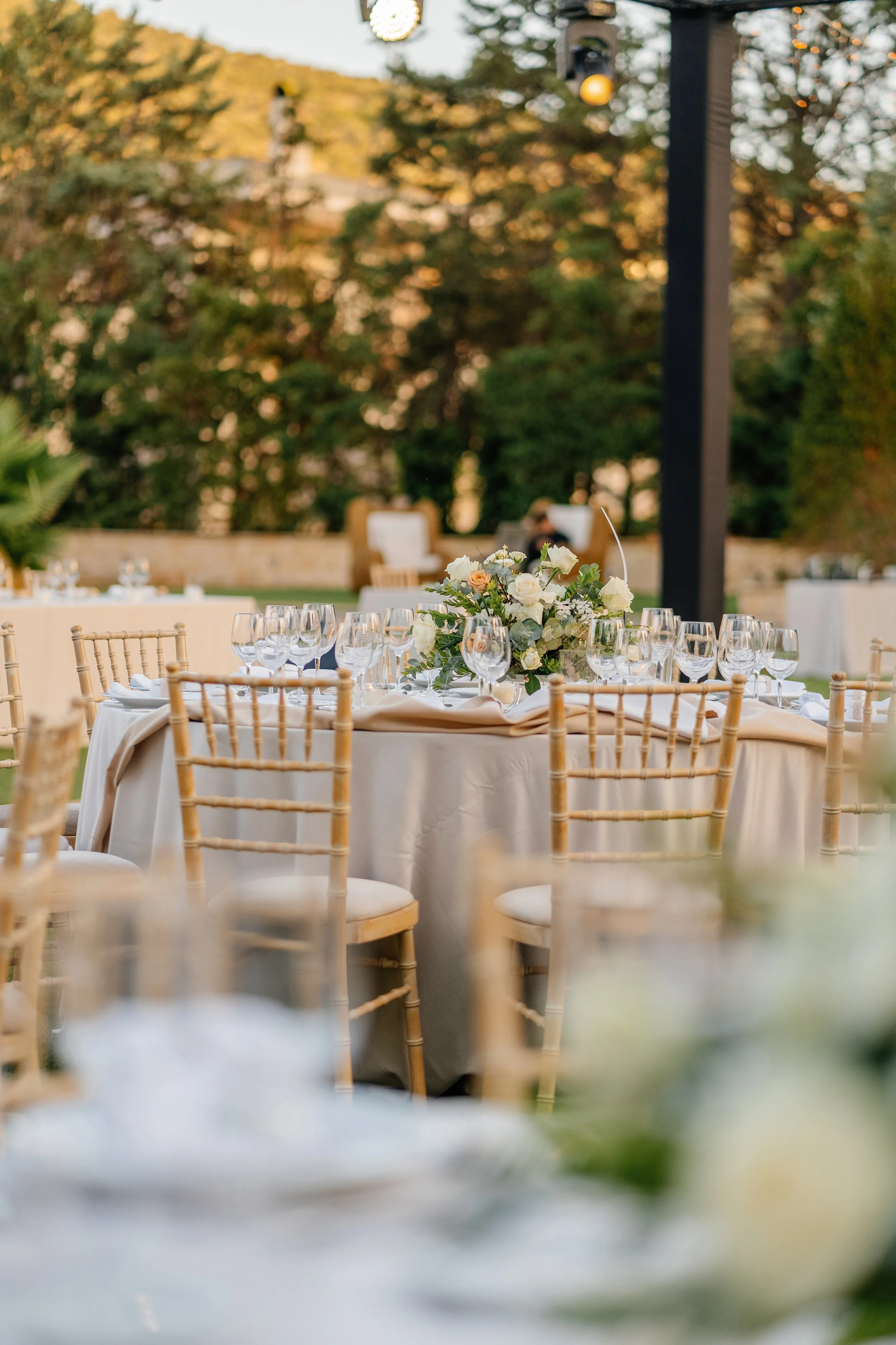 Set dining tables with beige chiavari chairs, white tablecloths, glassware, and a floral centerpiece with white and peach flowers, outdoors during daytime with trees and hanging lights in the background.