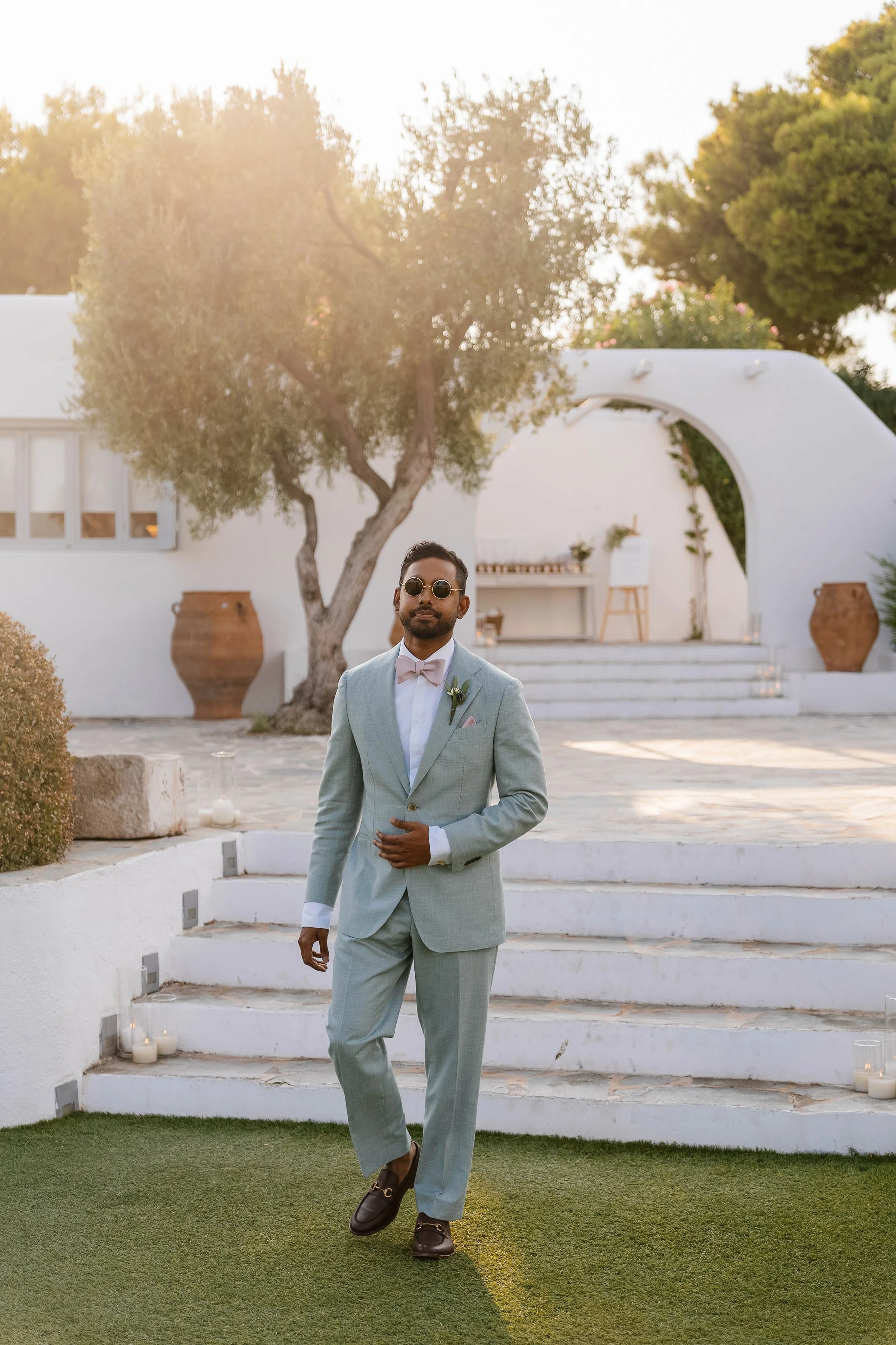 A man dressed in a light grey suit with sunglasses standing outdoors on a white staircase, with greenery and a white archway structure in the background during sunset.