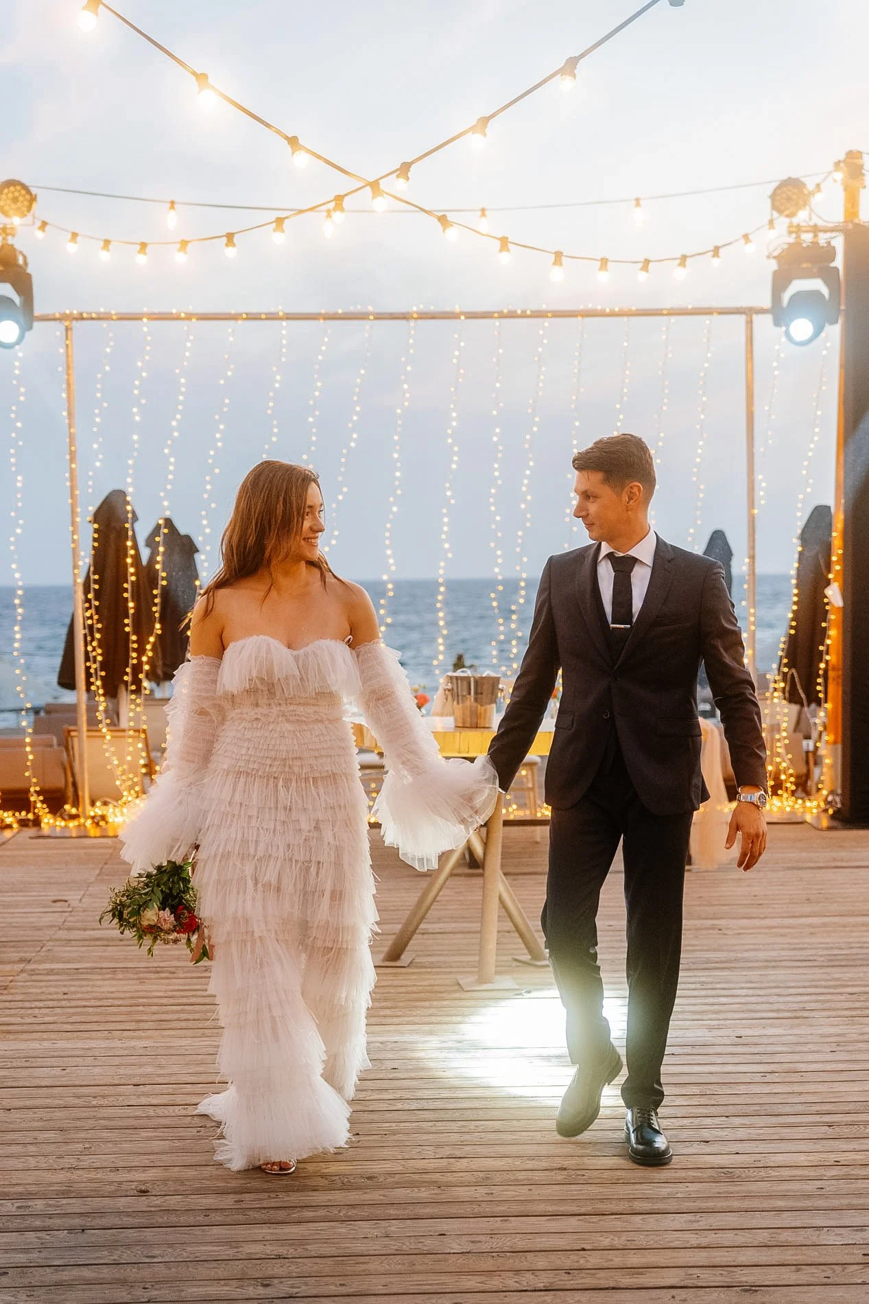 A newlywed couple walking hand in hand during their wedding reception by the sea at sunset, with string lights hanging overhead and string lights in the background.