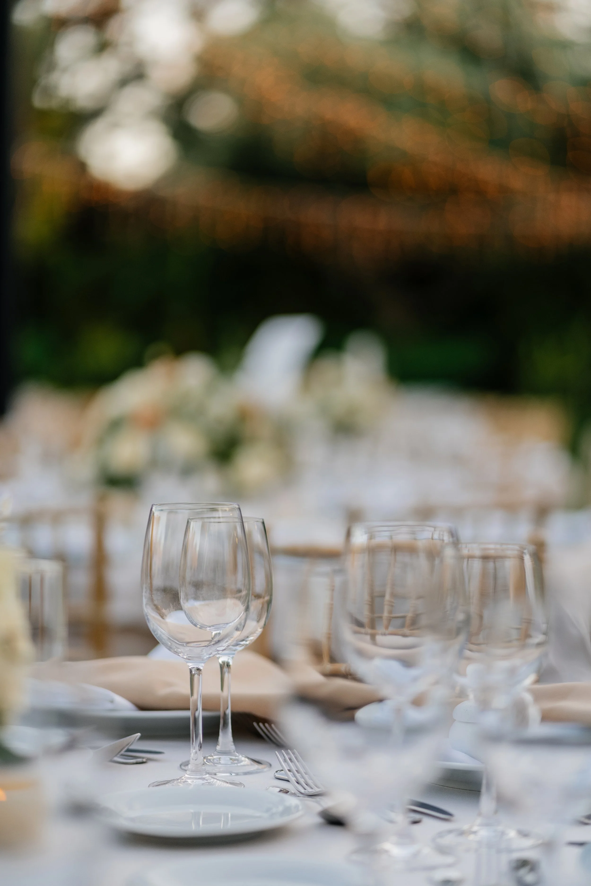 Elegant table setting with wine glasses, plates, silverware, and floral centerpieces, with a blurred outdoor background.