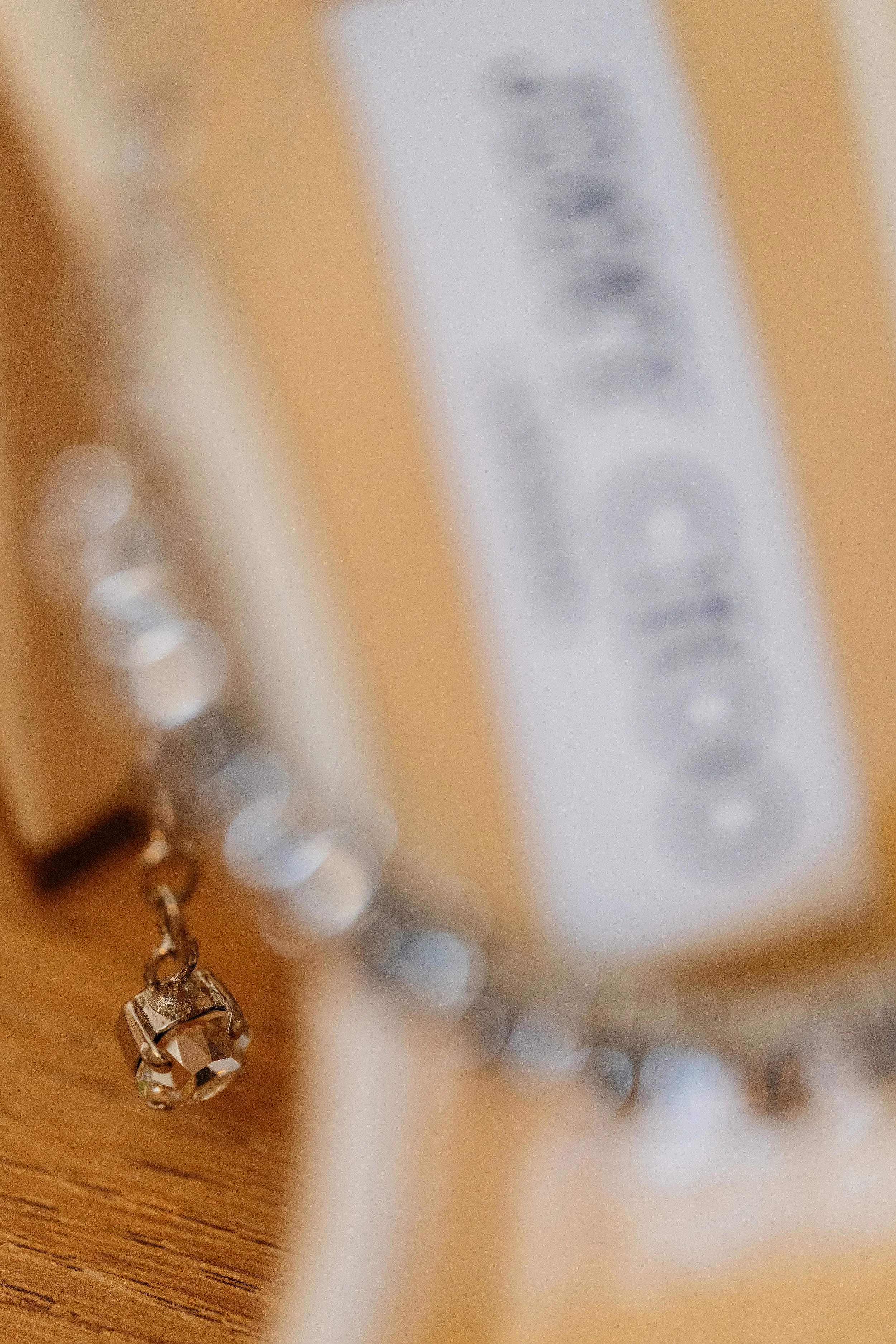 Close-up of a silver bracelet with a large, clear gemstone on a wooden surface, with a blurred white identification tag in the background.