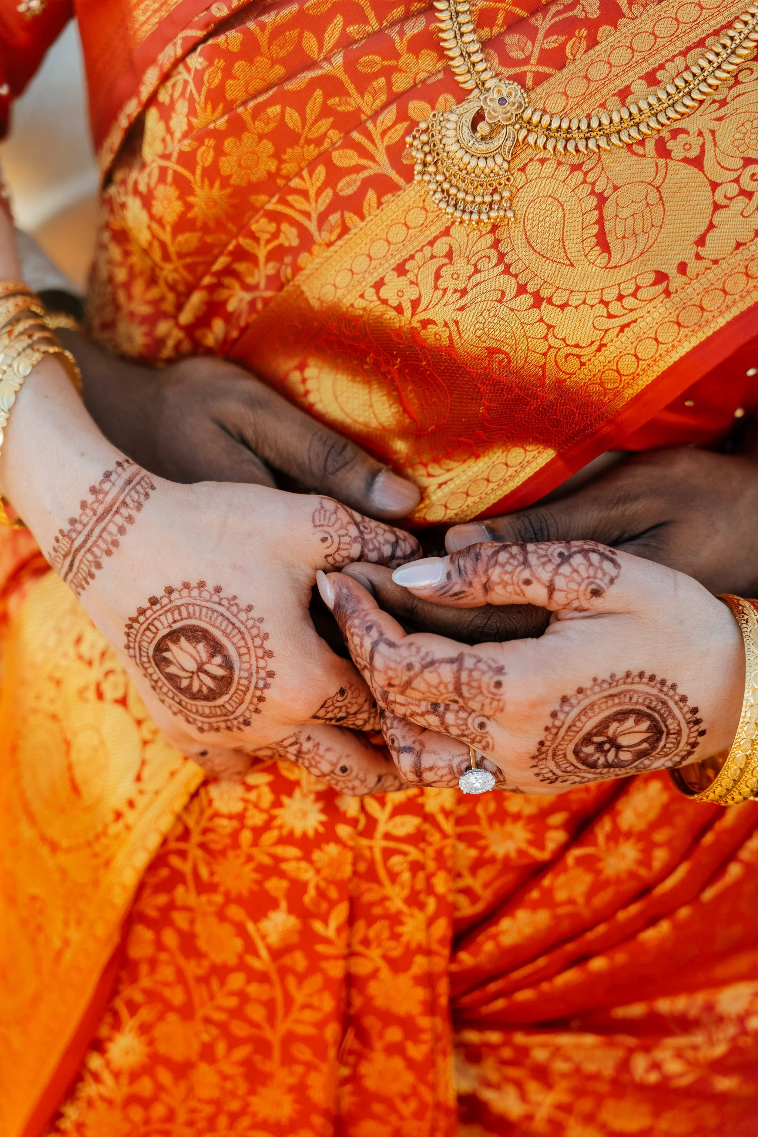 Close-up of a woman wearing an orange saree with intricate gold patterns, adorned with gold jewelry, including a necklace and bangles. Her hands and wrist are decorated with mehndi (henna) designs, and she is wearing a diamond ring.
