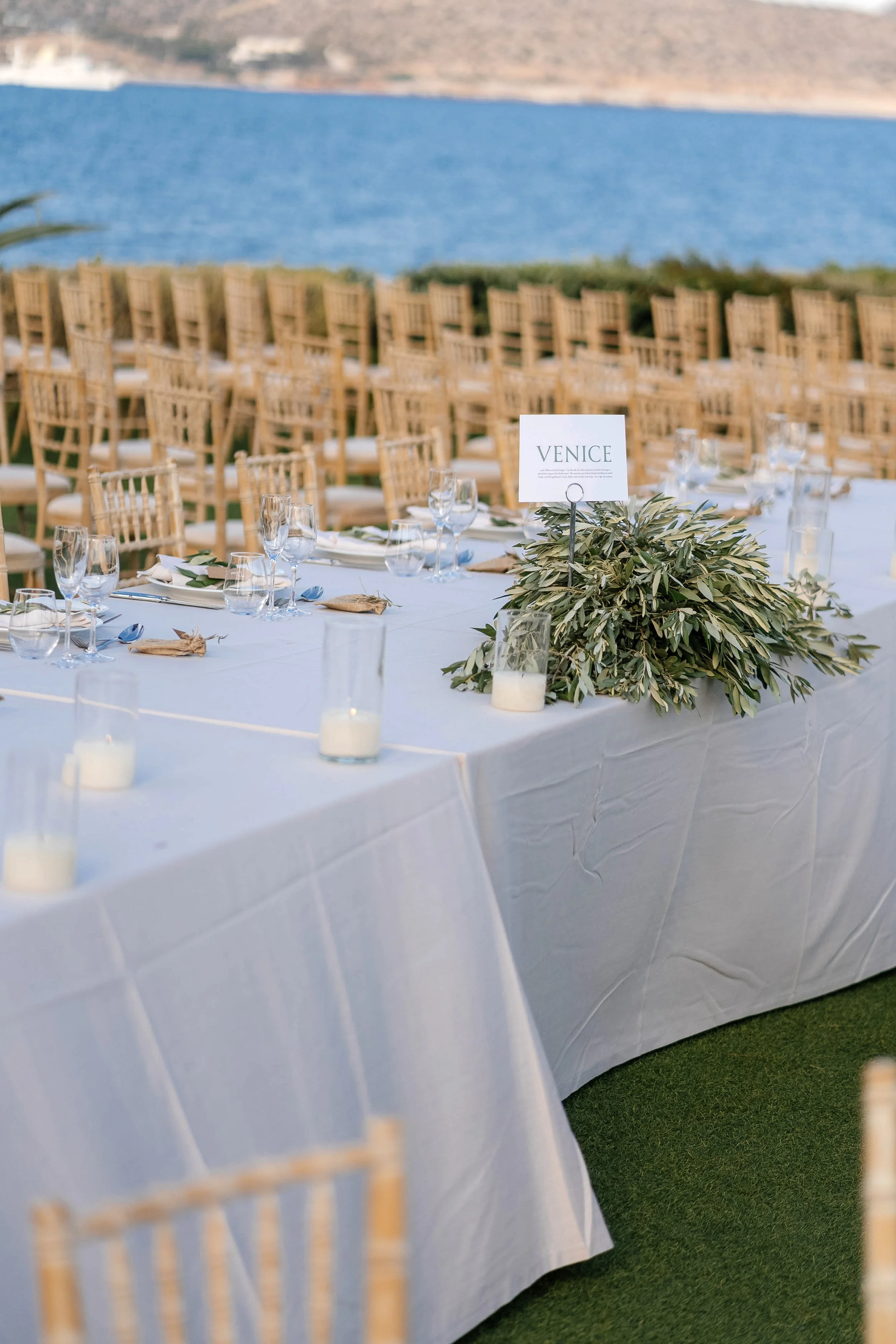 Outdoor wedding reception setup by a lake with a long white table, multiple chairs, glassware, candles, and a centerpiece with greenery and a 'Venice' sign.