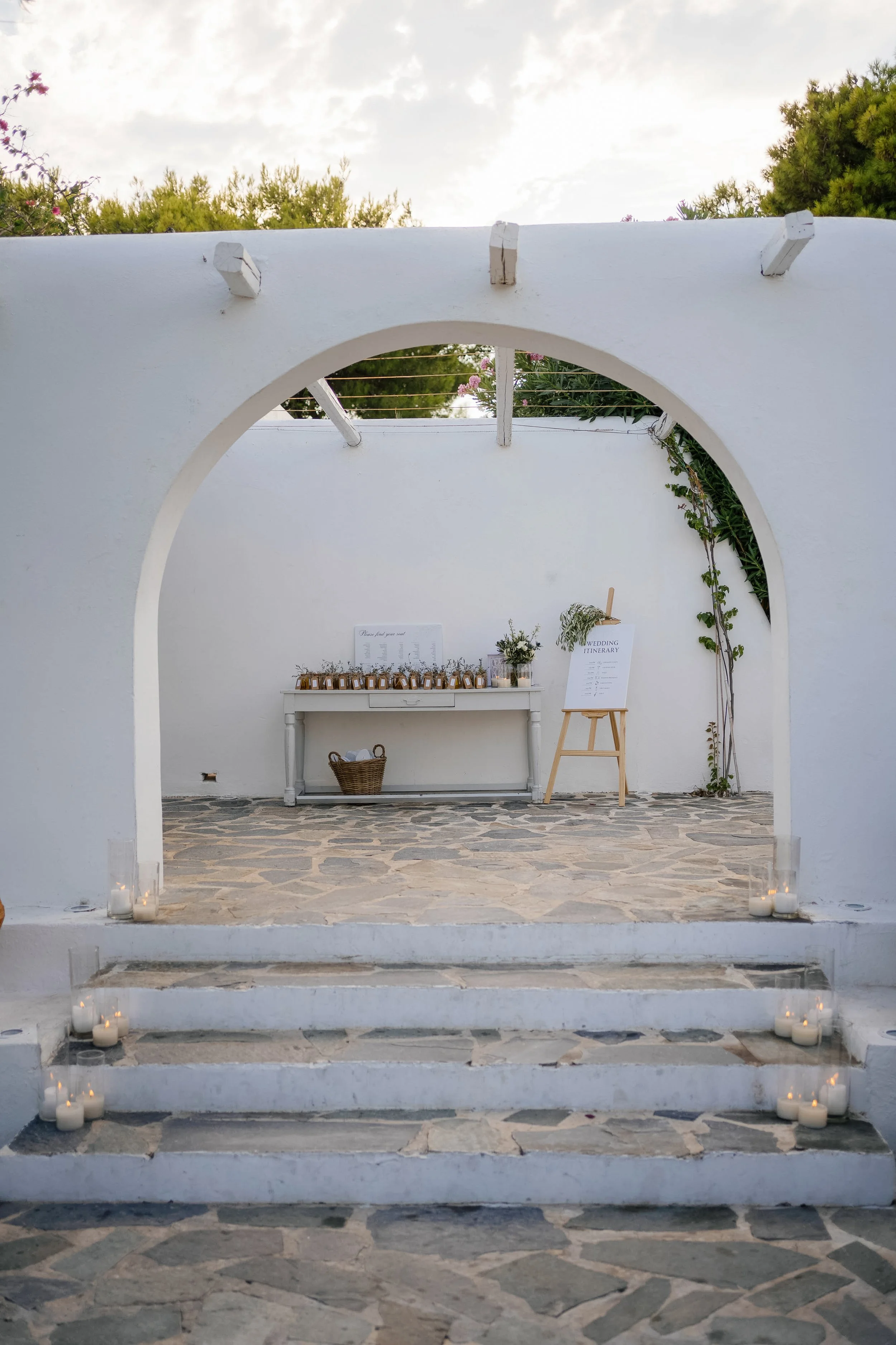 A white archway leading to an outdoor wedding or event area, with small candles in glass holders on the ground and a white table with floral arrangements and a wedding itinerary sign.