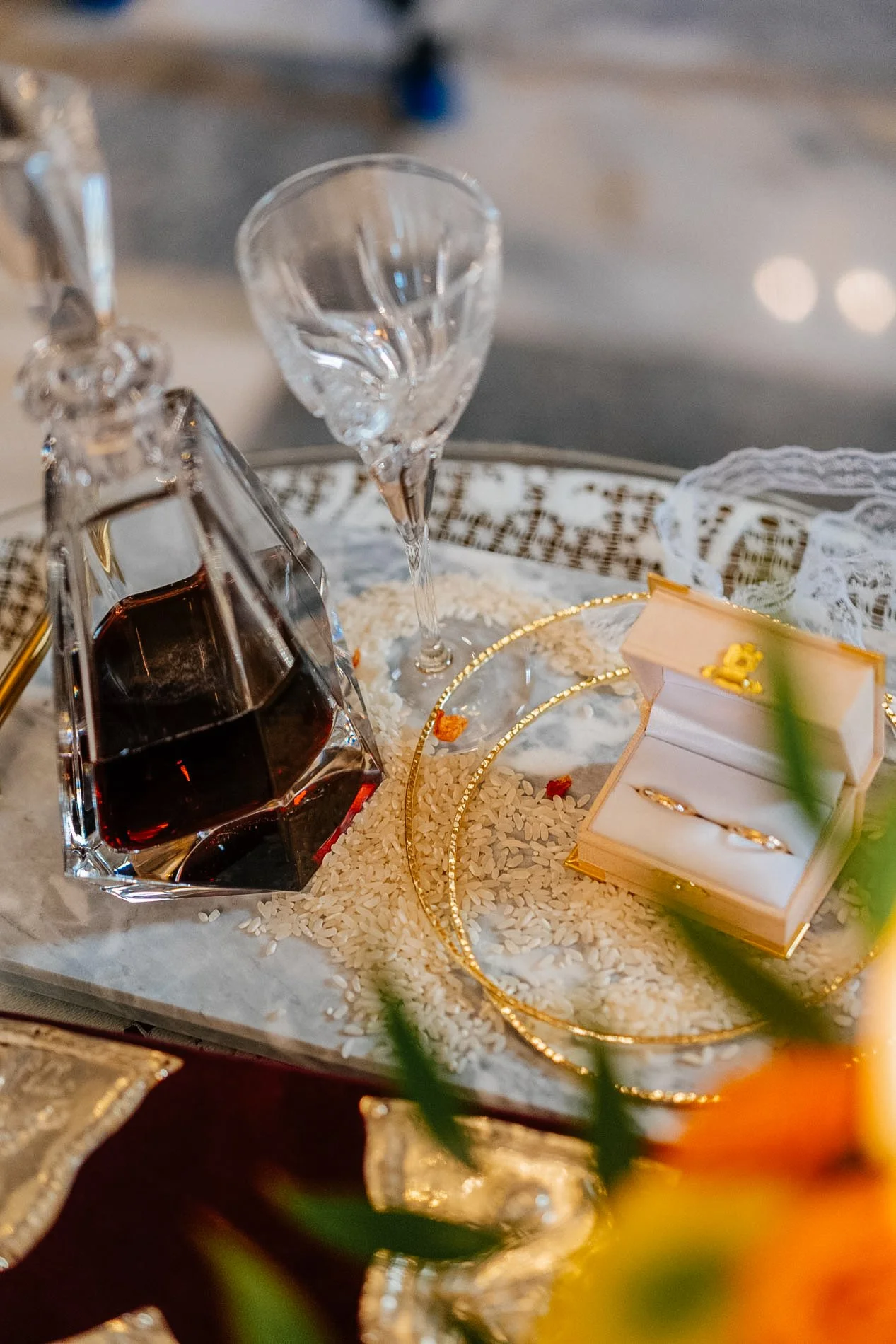 A decorative display featuring a pair of gold wedding rings in a white jewelry box, a glass of wine, a decanter of red wine, and a small open box with more rings, all arranged on a tray with scattered rice and gold accents.