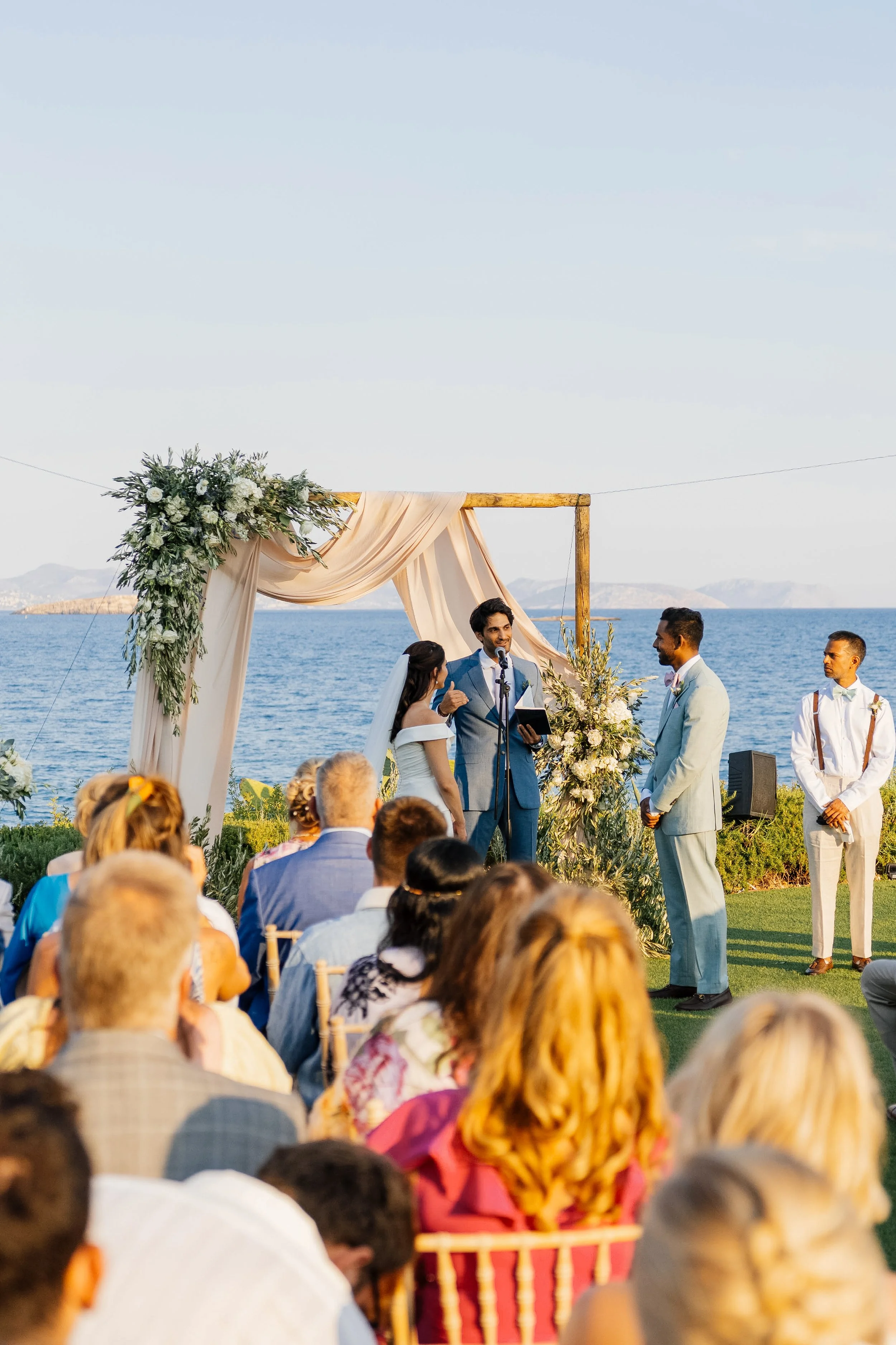 A wedding ceremony taking place outdoors by the water, with a decorated archway, a wedding officiant, the bride and groom, and guests seated facing the couple.