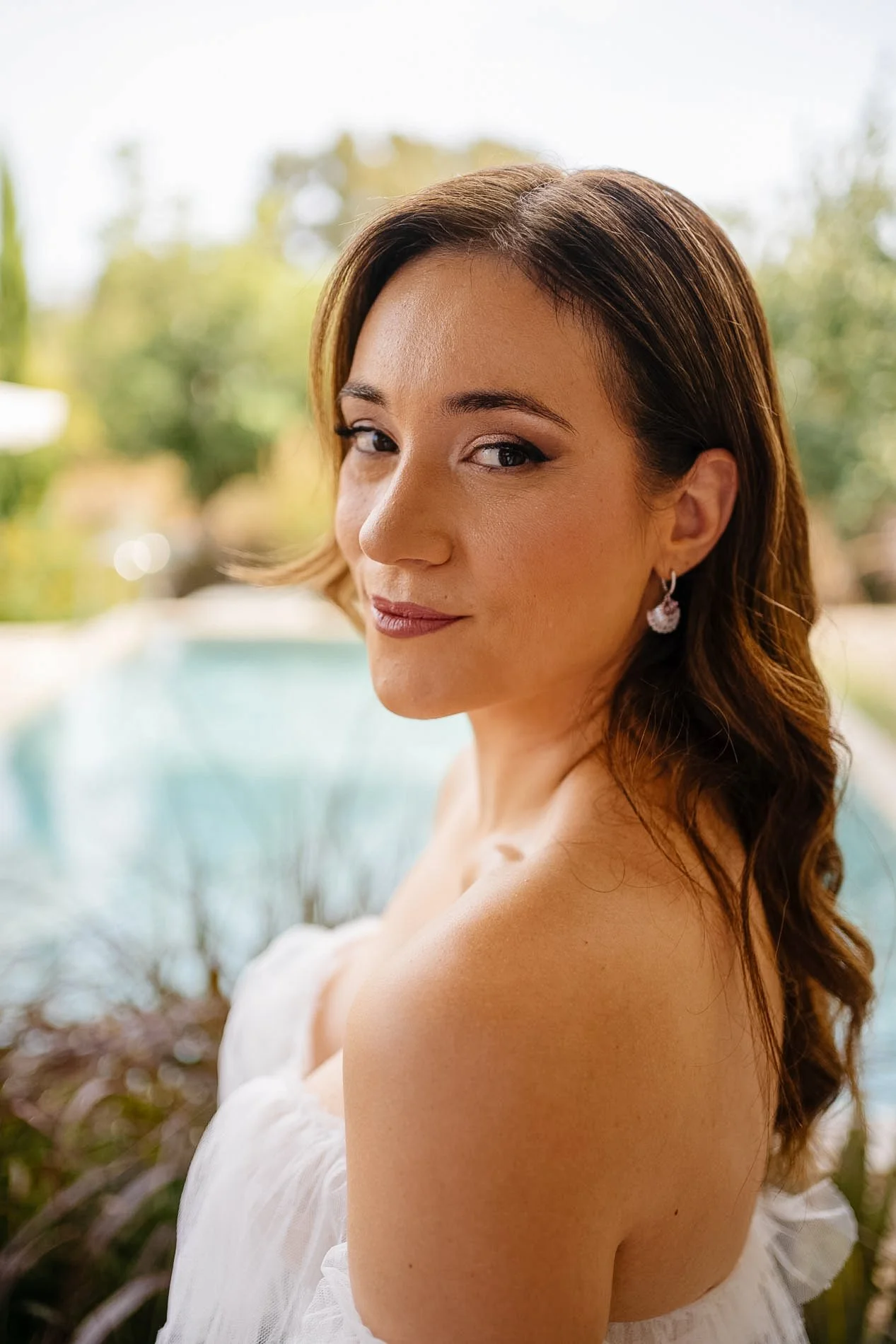 A woman with long, wavy brown hair and earrings, wearing a strapless white dress, standing outdoors with a blurred background of trees and water.