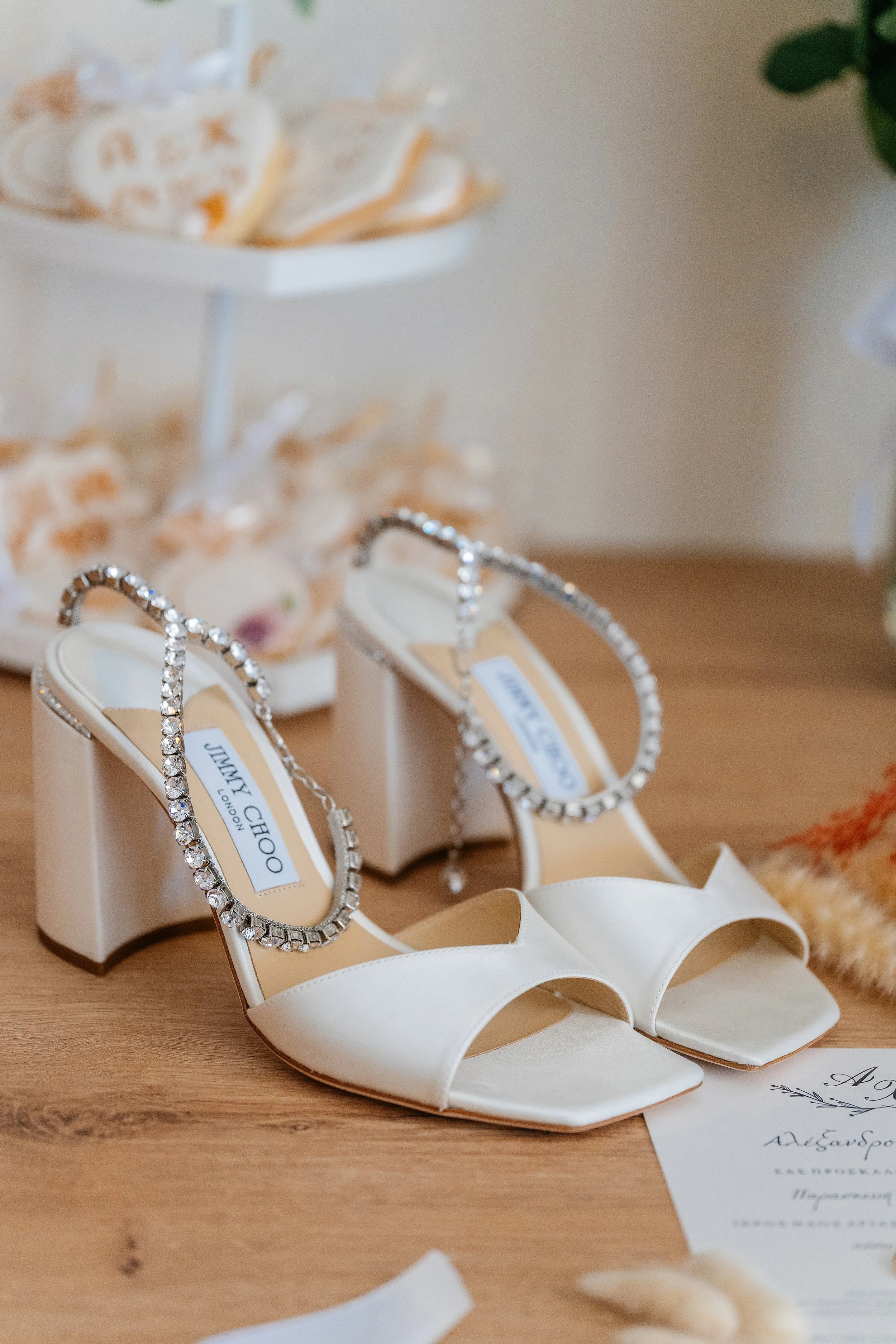 White high-heeled shoes with rhinestone details displayed on a wooden surface, with a white box and a paper, possibly for a wedding or special event.
