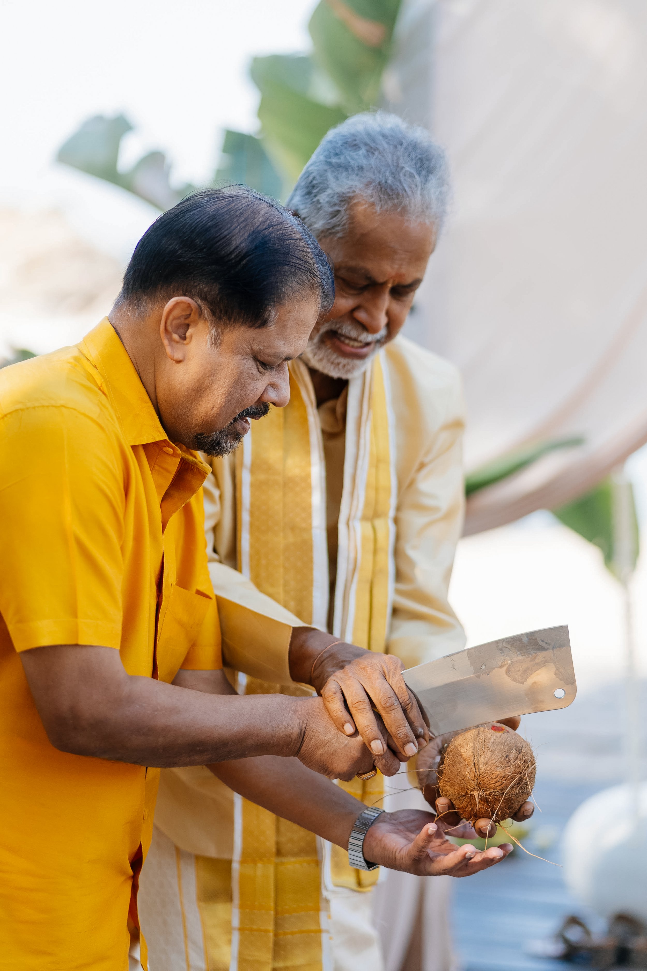 Two men in yellow shirts examining a coconut with a machete outdoors.