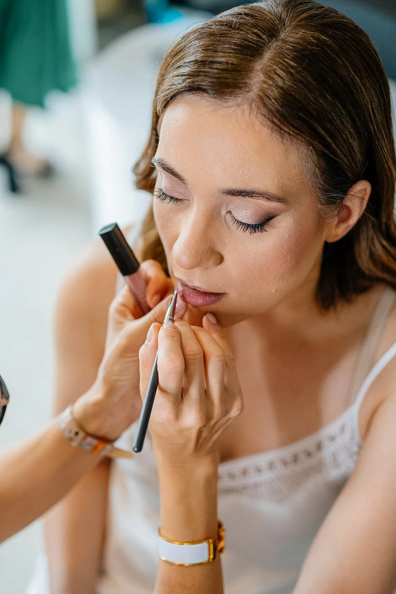 Close-up of a woman with brown hair and light skin receiving lip makeup application from a makeup artist, with her eyes closed.
