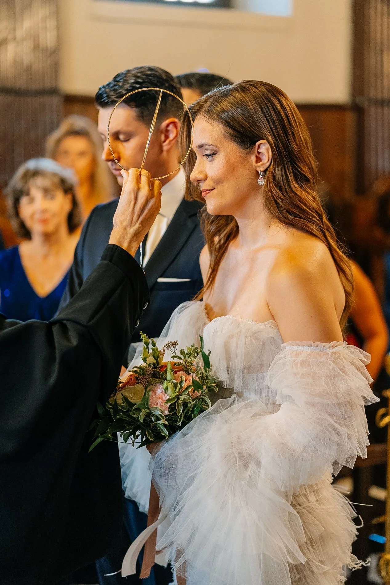 A bride in a white wedding dress holding a bouquet of flowers during a wedding ceremony, with an officiant holding a jewelry ring over her head.