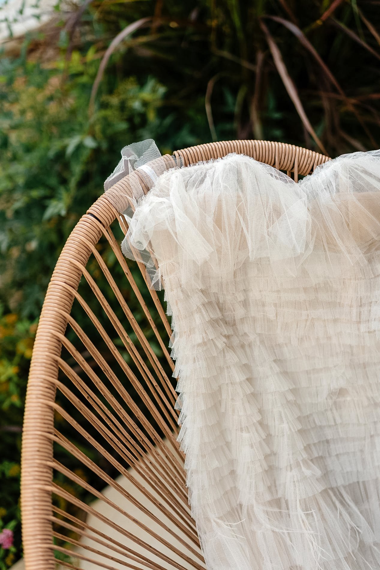 Close-up of a white, ruffled dress with tulle fabric draped over the edge of a wicker chair, outdoors with greenery in the background.