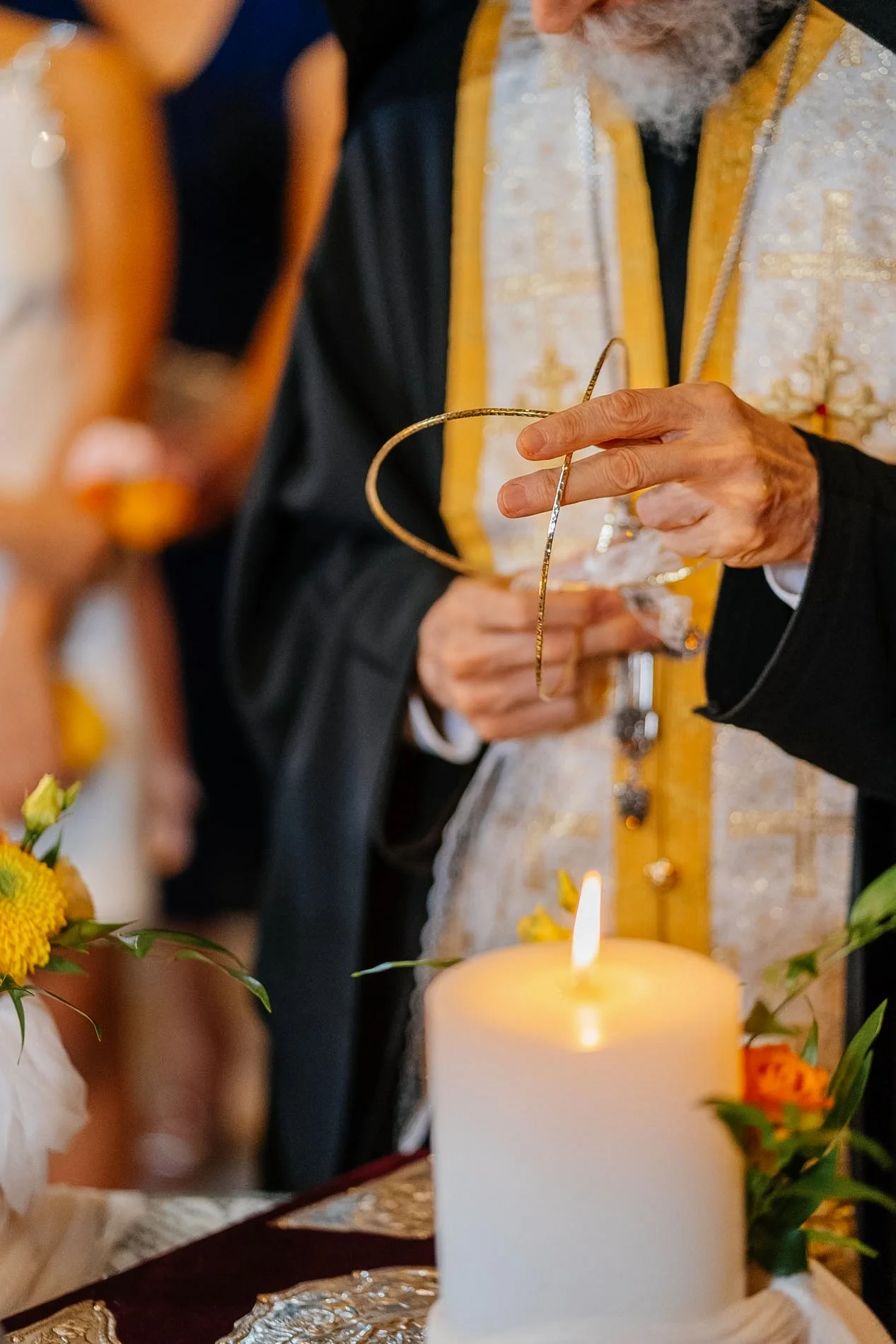 A priest holds a golden wedding ring over a lit candle during a wedding ceremony, with flowers and a decorated altar in the background.
