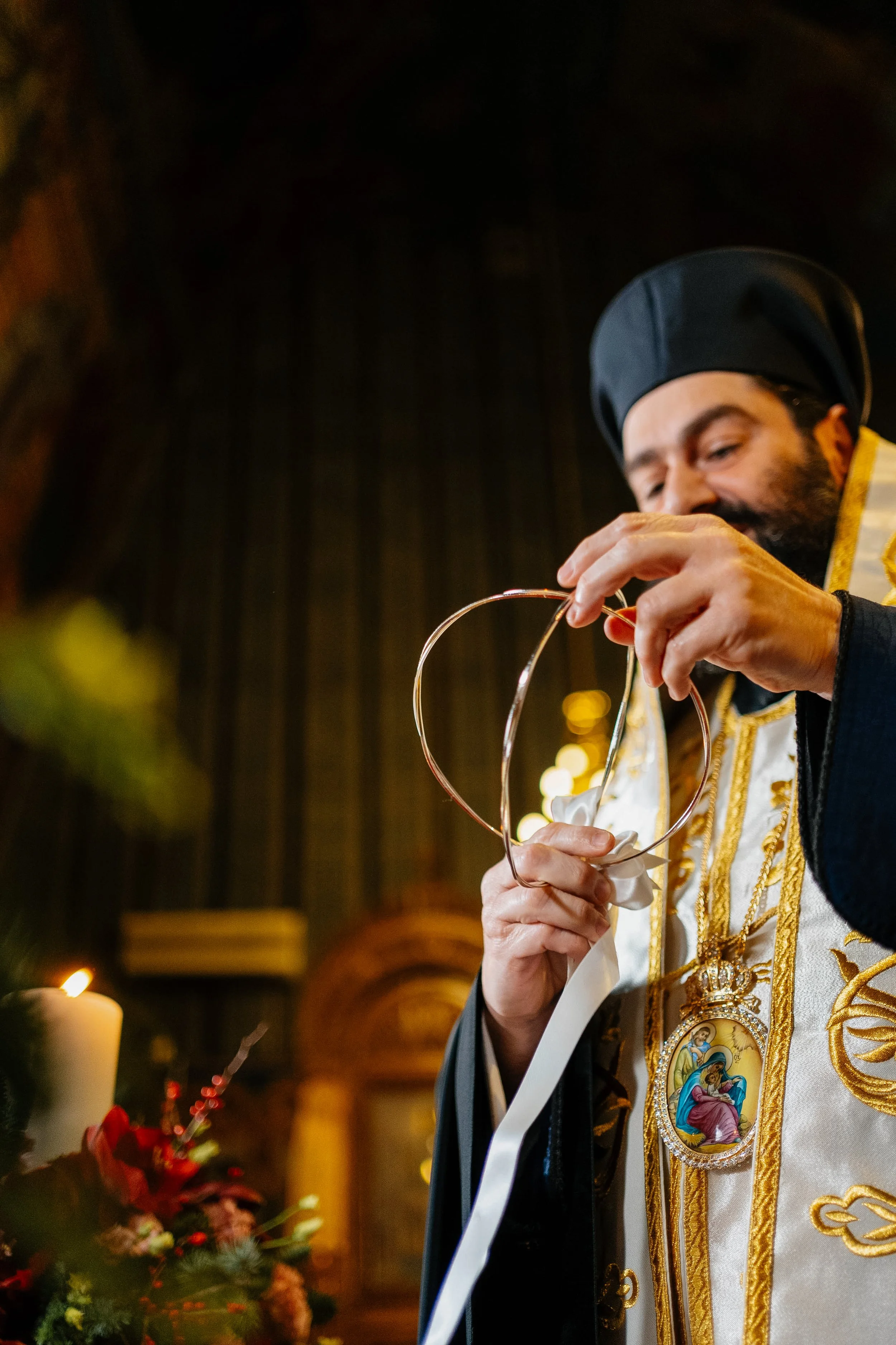 A priest dressed in ornate gold and white vestments with religious icons on the front, performing a ritual or blessing with a metal object in a dimly lit setting.