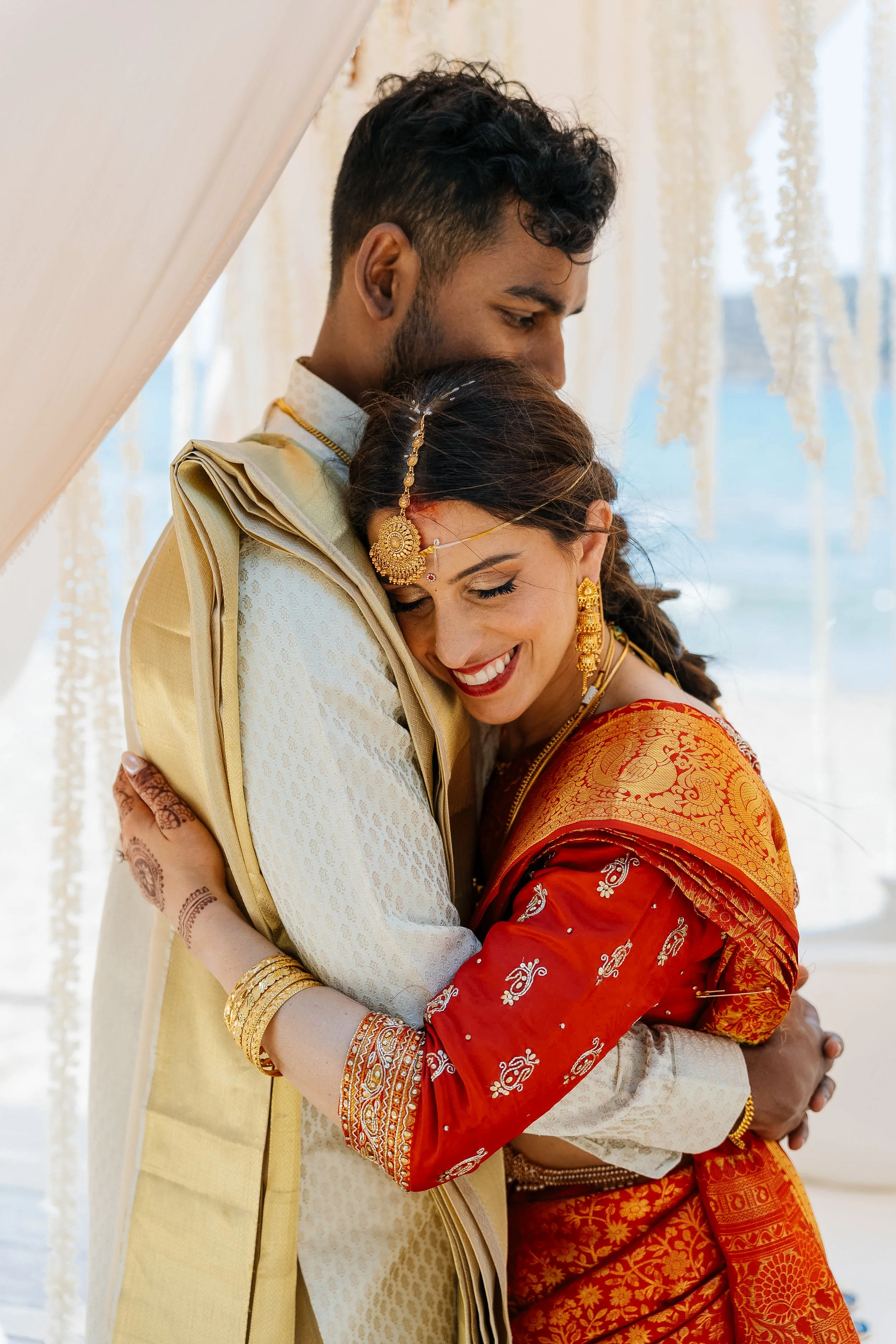 A newlywed Indian couple embraces, with the groom dressed in traditional cream and gold attire and the bride in a vibrant orange and gold saree, both adorned with jewelry, smiling with their eyes closed.