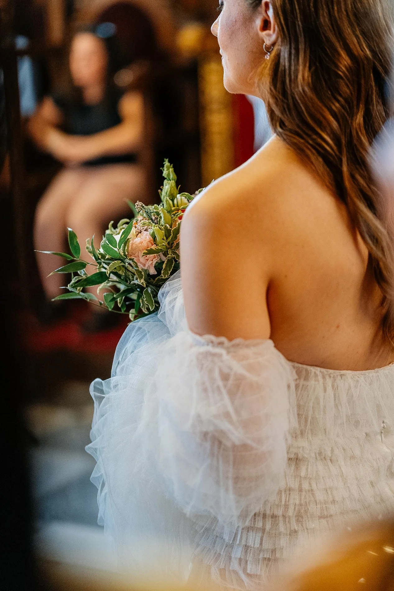 Woman holding a bouquet of flowers, wearing a white dress with sheer puffed sleeves, seen in profile, with other people blurred in the background.
