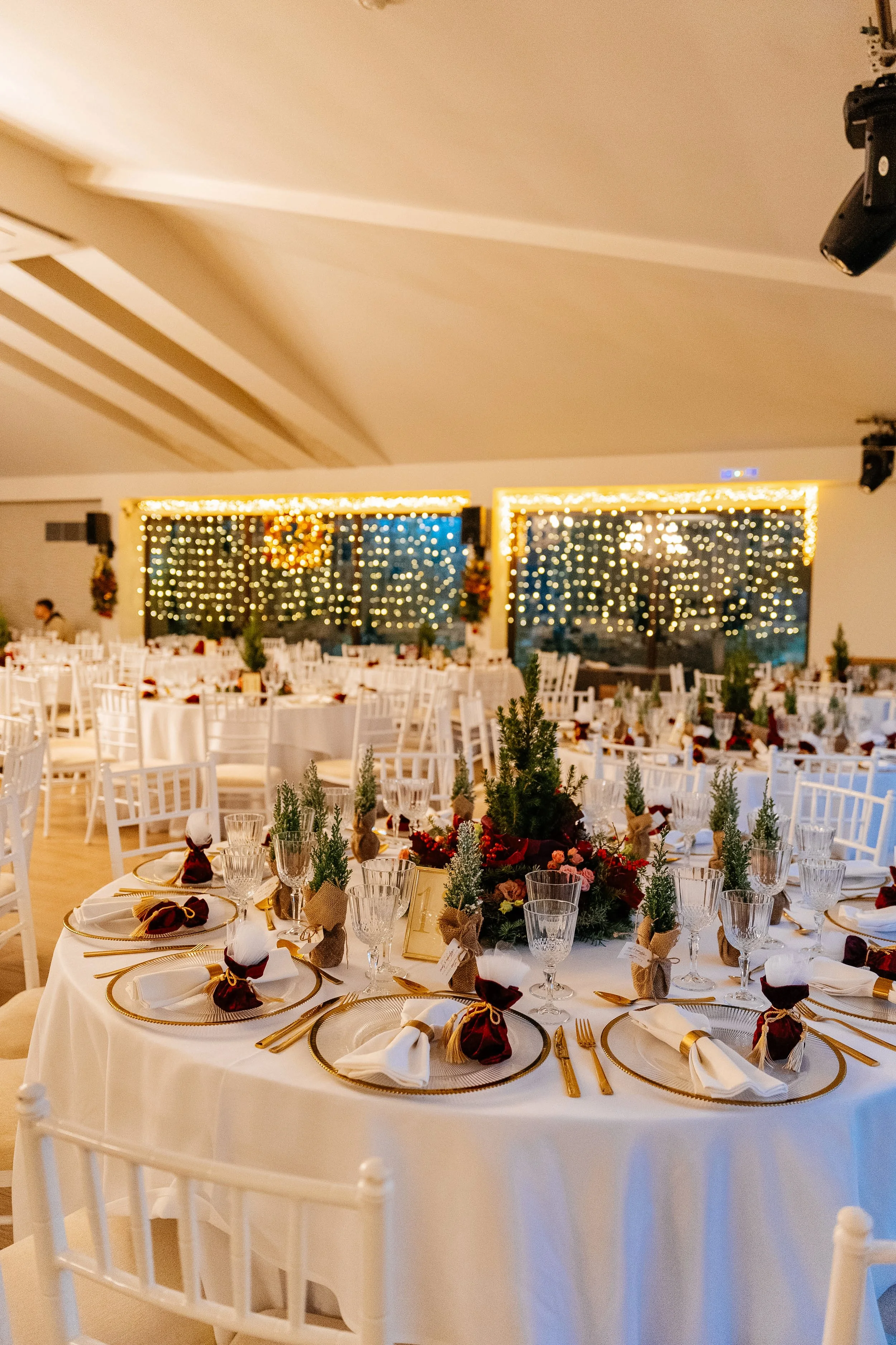 A decorated banquet hall with round tables, elegant place settings, and floral centerpieces. White chairs surround the tables, and there are string lights and decorative curtains with lights in the background.