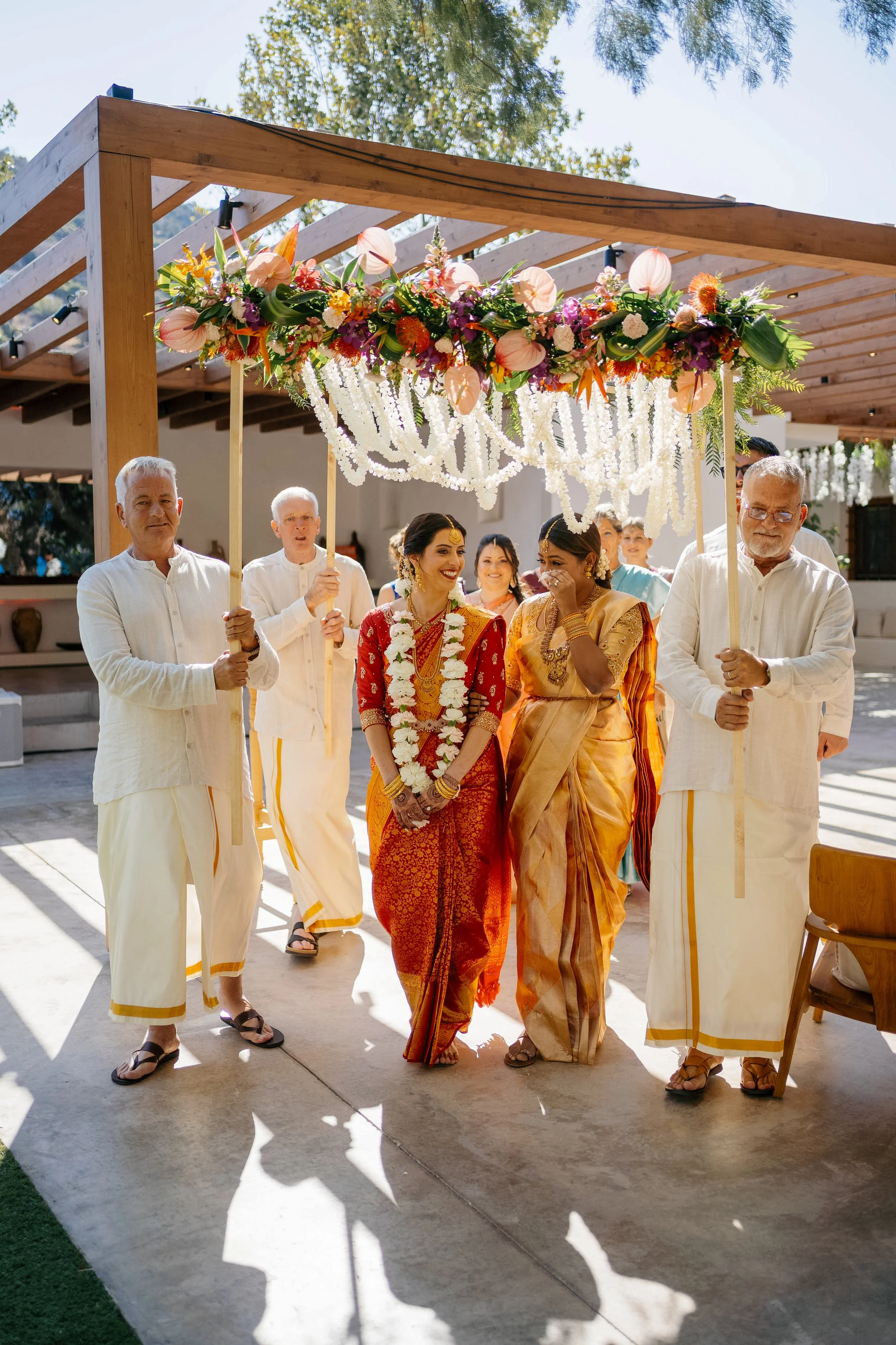 A group of people of different ethnicities, including a woman in traditional Indian attire, walking under a wooden decorated canopy at a wedding or celebration. The canopy is adorned with colorful flowers and white floral strands, and the people appe