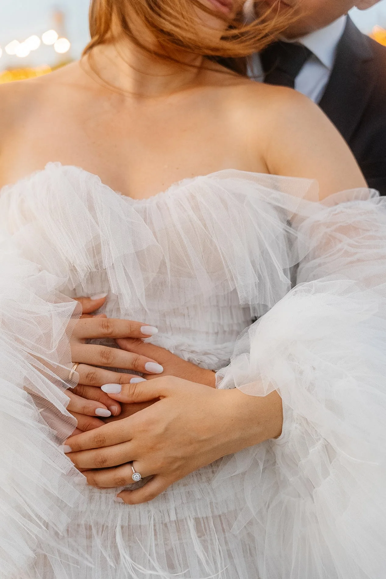 Close-up of a bride in a white dress with ruffle details, holding her baby bump, with a ring on her finger. A man in a suit is partially visible behind her.