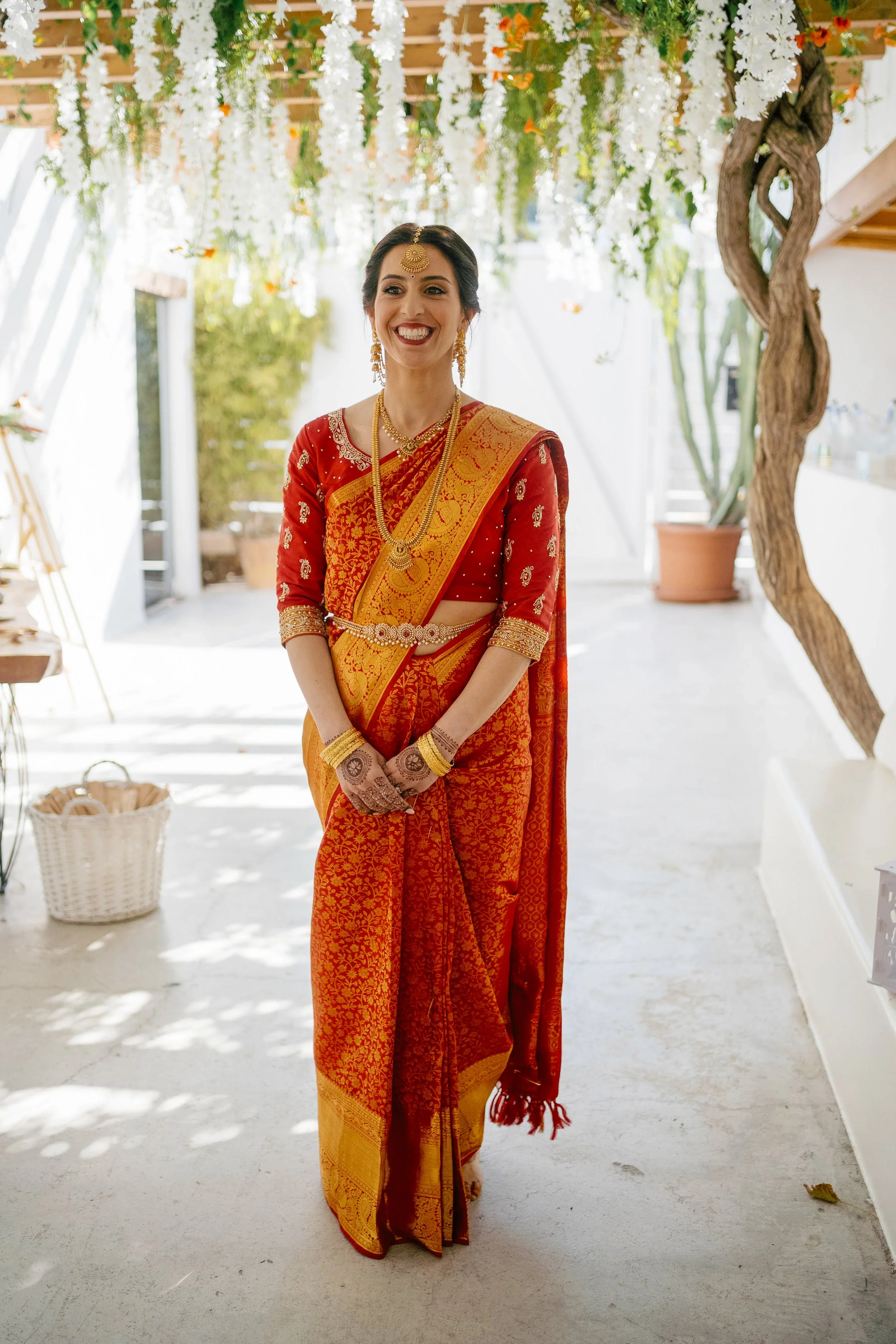 A woman dressed in traditional Indian attire, wearing a red and gold sari, jewelry, and mehndi on her hands, standing indoors with floral decorations hanging above.