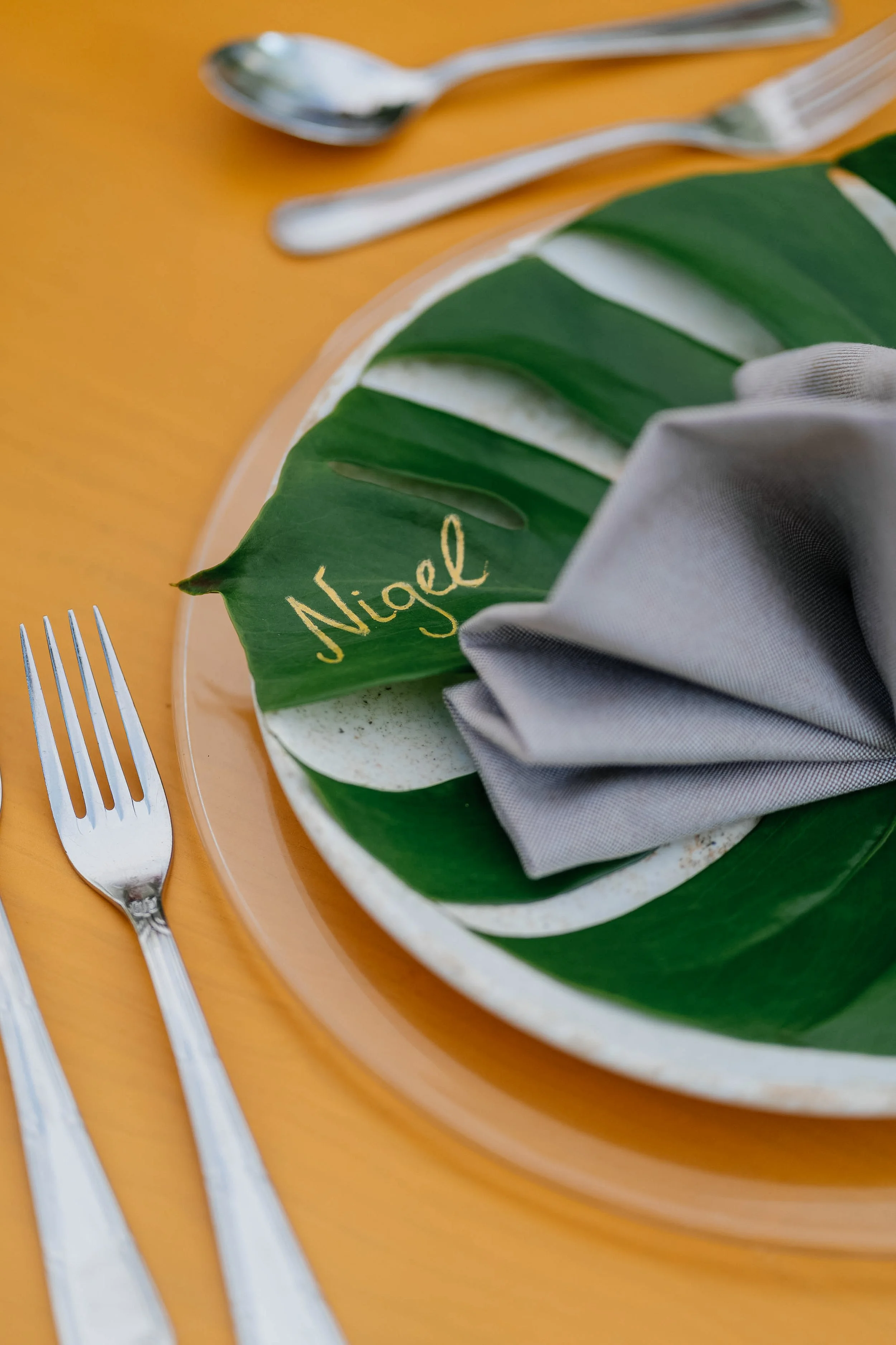 A close-up of a table setting featuring a green leaf with the name 'Nigel' written in gold, a gray cloth napkin, and silverware on a yellow surface.
