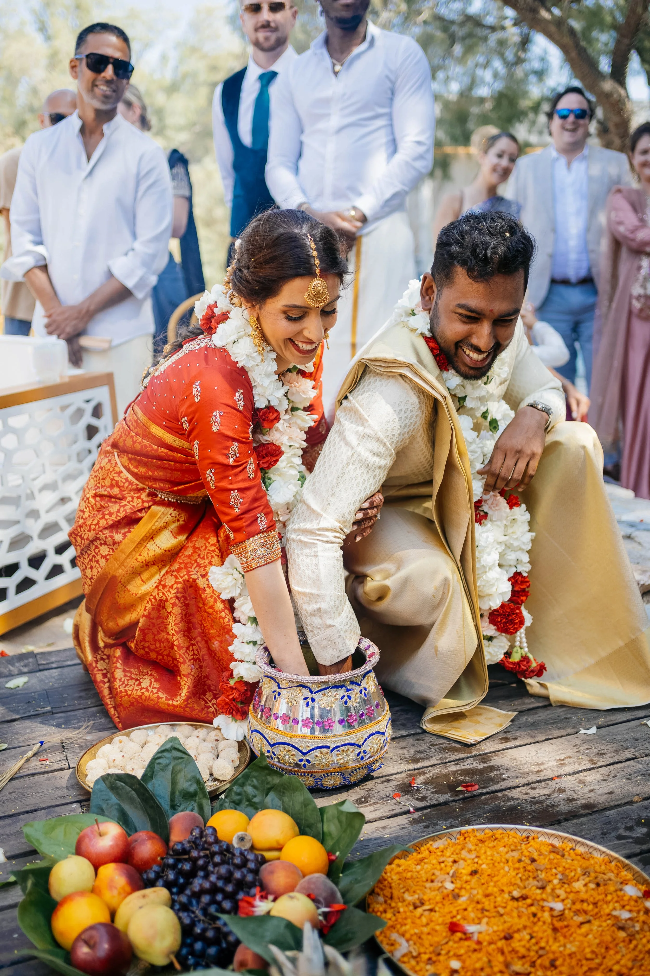 Indian wedding ceremony with bride and groom offering prayers, surrounded by friends and family, with fruits and traditional offerings in the foreground.