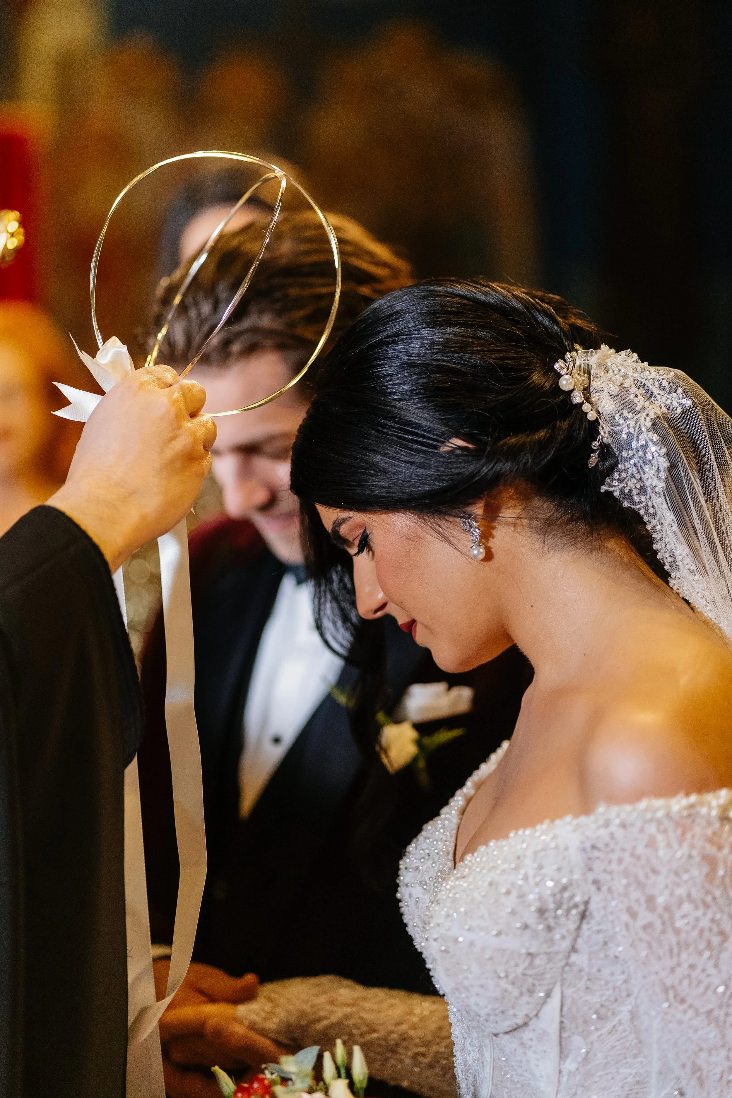 A bride and groom bowing their heads during a wedding ceremony, with a person holding a decorative metal hoop and ribbon in the foreground.
