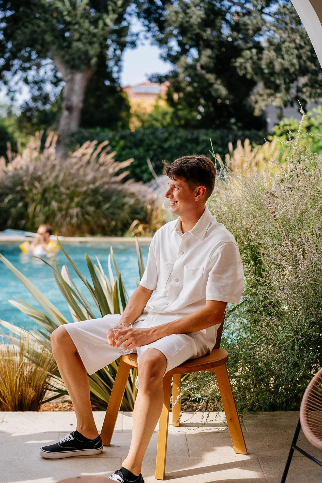 A young man sitting on a wooden stool outdoors by a swimming pool, smiling and holding a glass, surrounded by greenery and plants.