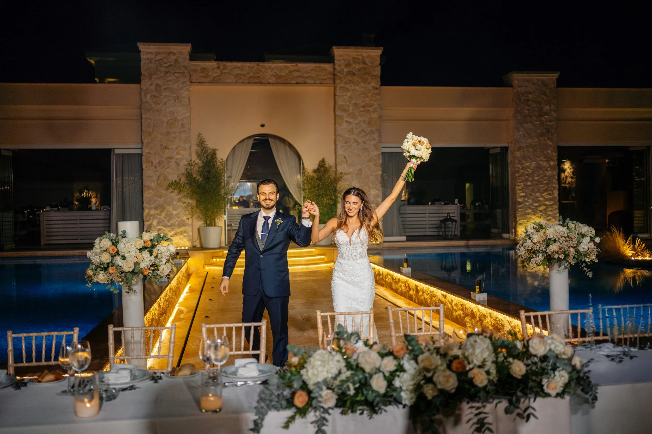 A bride and groom holding hands and smiling, celebrating at night on a decorated outdoor wedding pathway by a pool, with table settings and floral arrangements in the foreground.