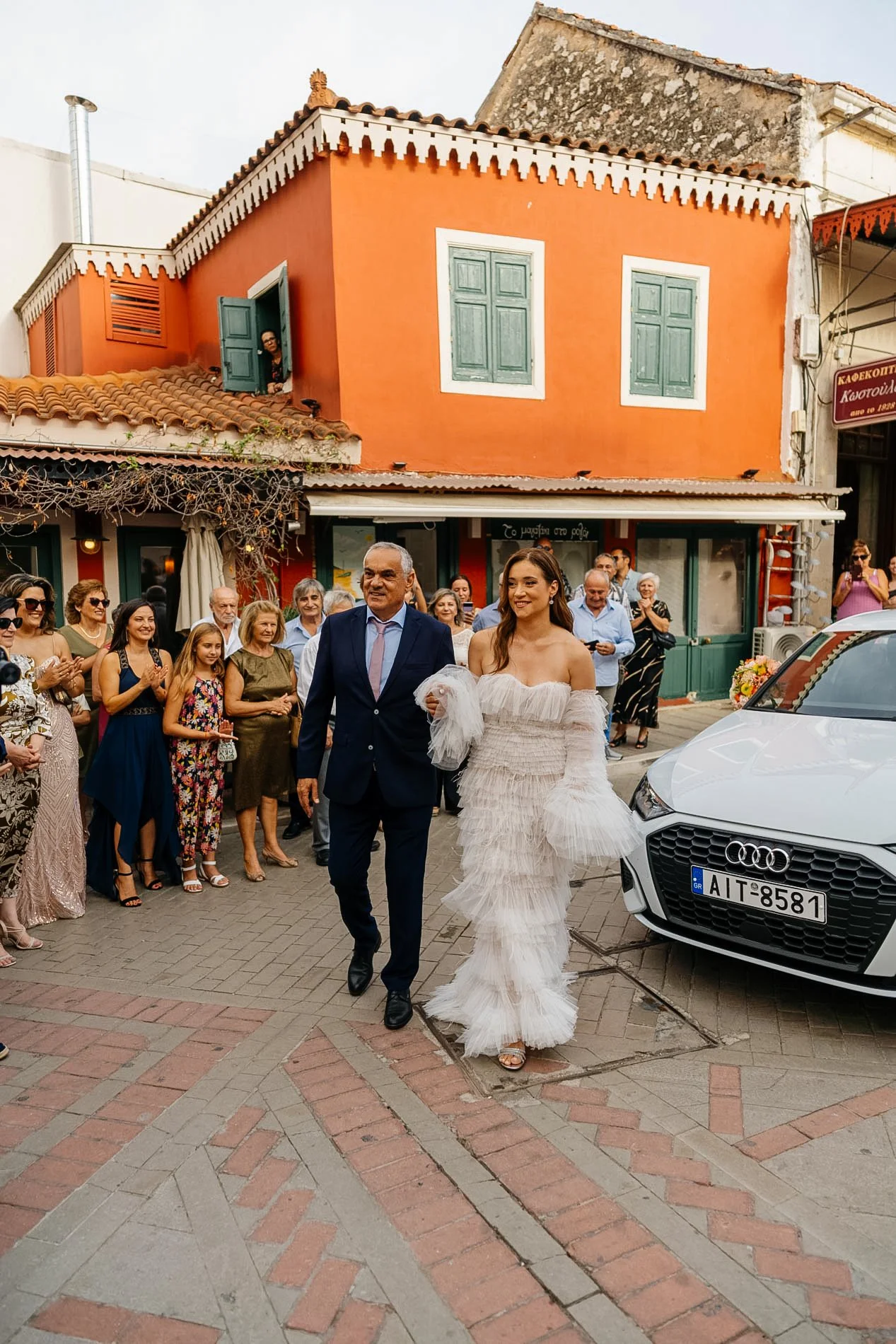 A bride in a white off-shoulder wedding dress smiling as she walks with an older man, likely her father, on her wedding day. They are surrounded by guests outside a colorful building painted in orange and white, with a white Audi car parked nearby.