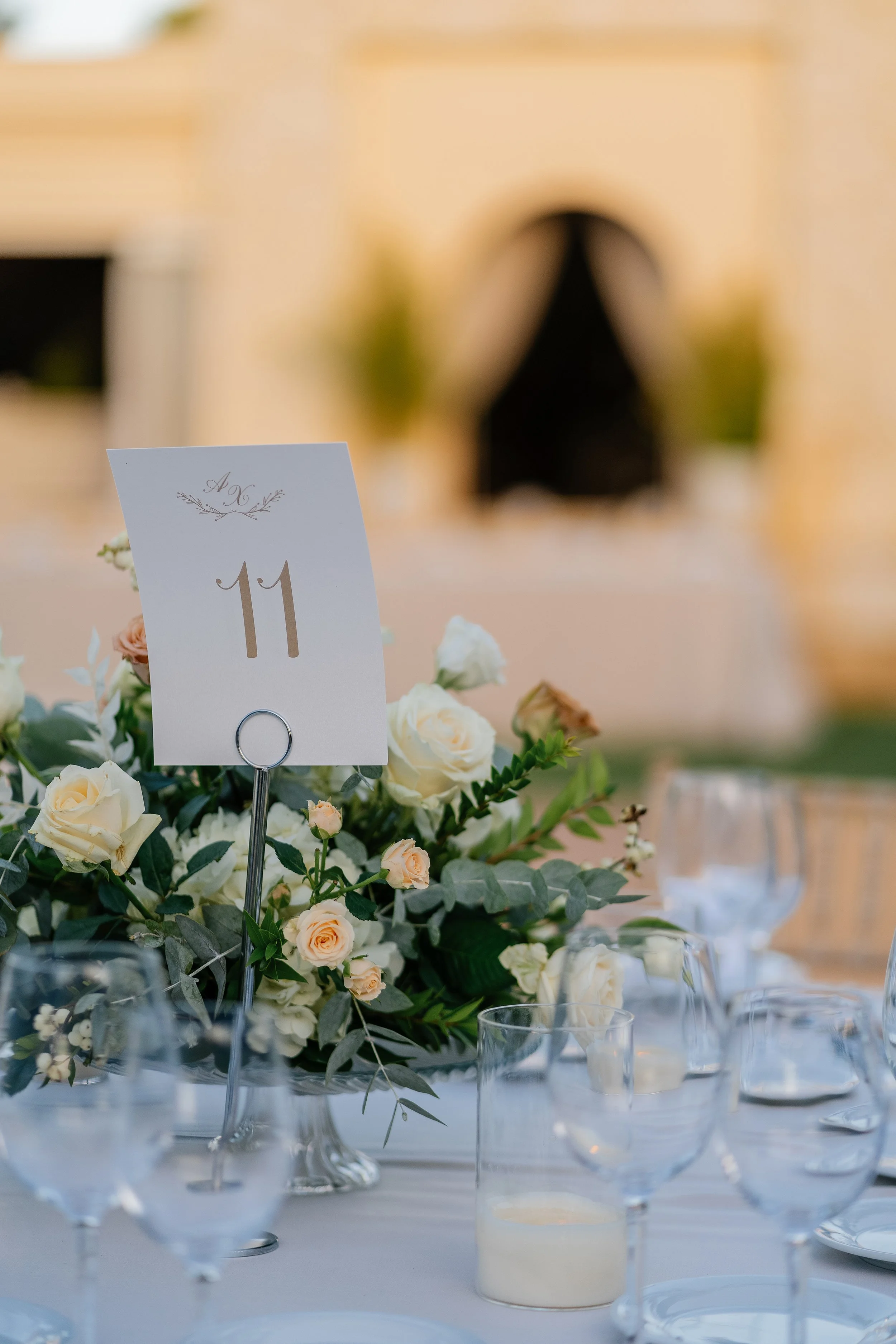 Table centerpiece with white and light peach roses, greenery, and a table number card '11'. The table is decorated with glassware and candles, and there is a blurred indoor background.