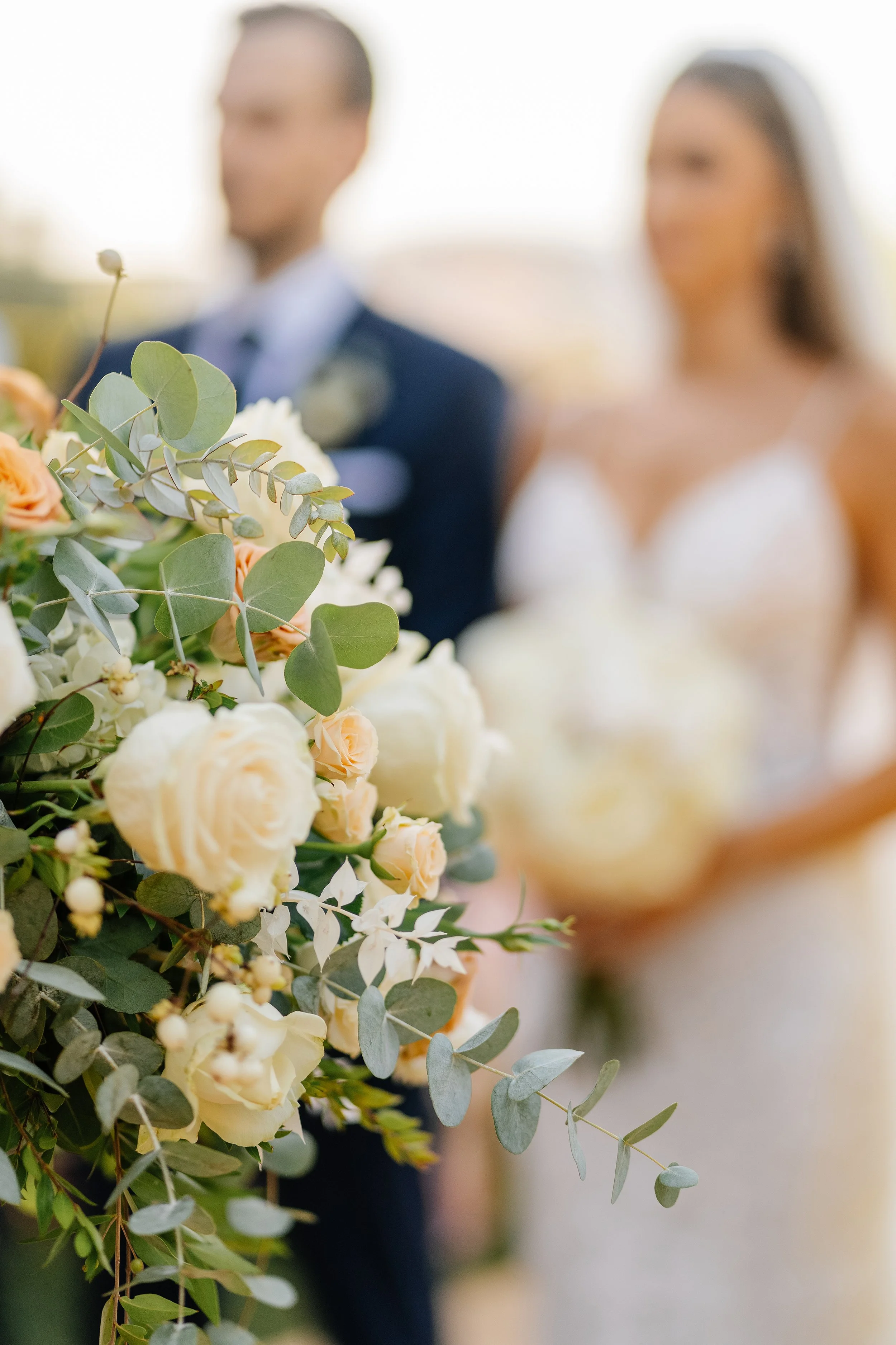 A blurred couple standing in wedding attire behind a large bouquet of white and peach roses with greenery, outdoors on a sunny day.