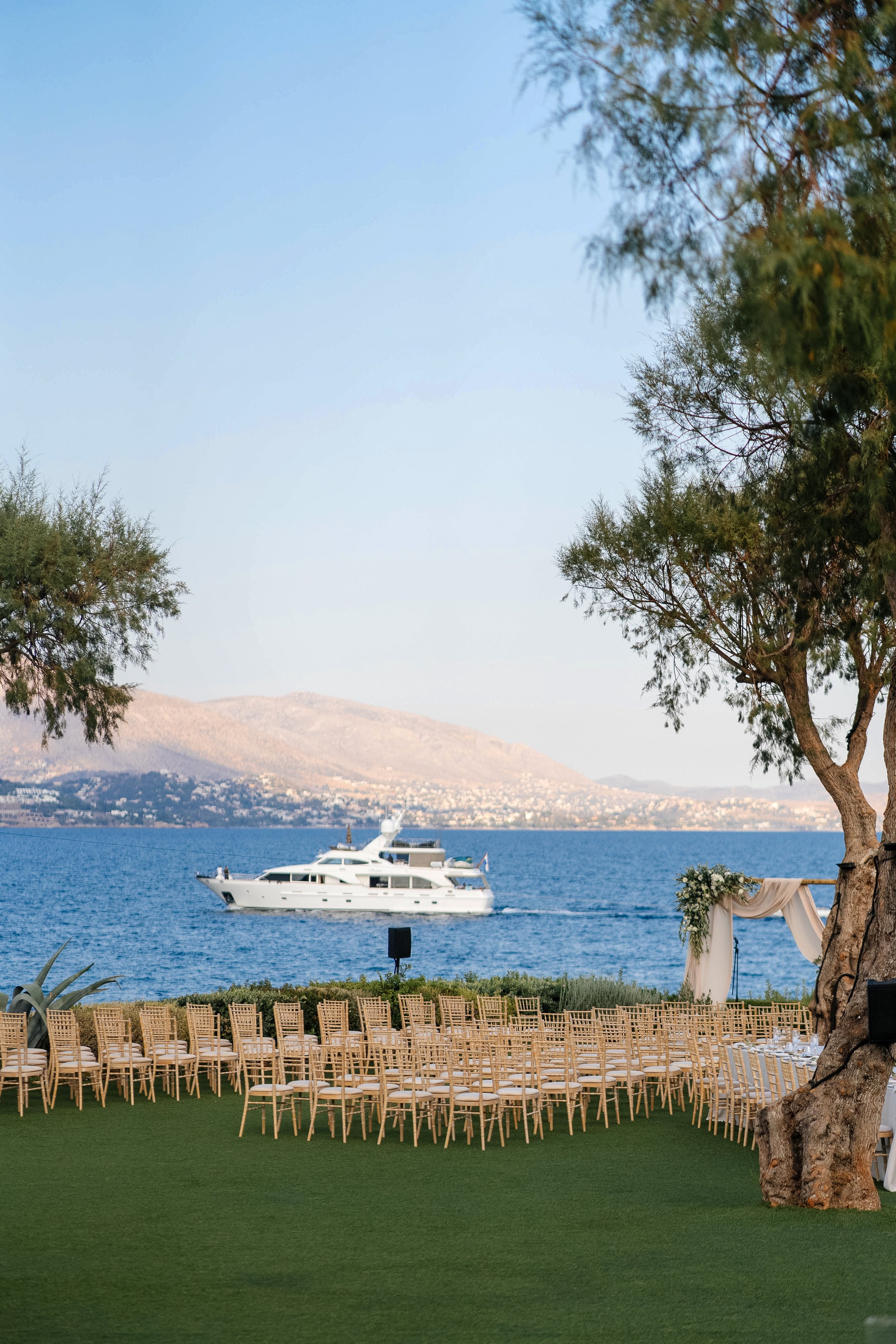 Yacht sailing on a lake with mountains in the background, set up for an outdoor event with chairs and an arch draped with cloth and flowers. Trees frame the scene.