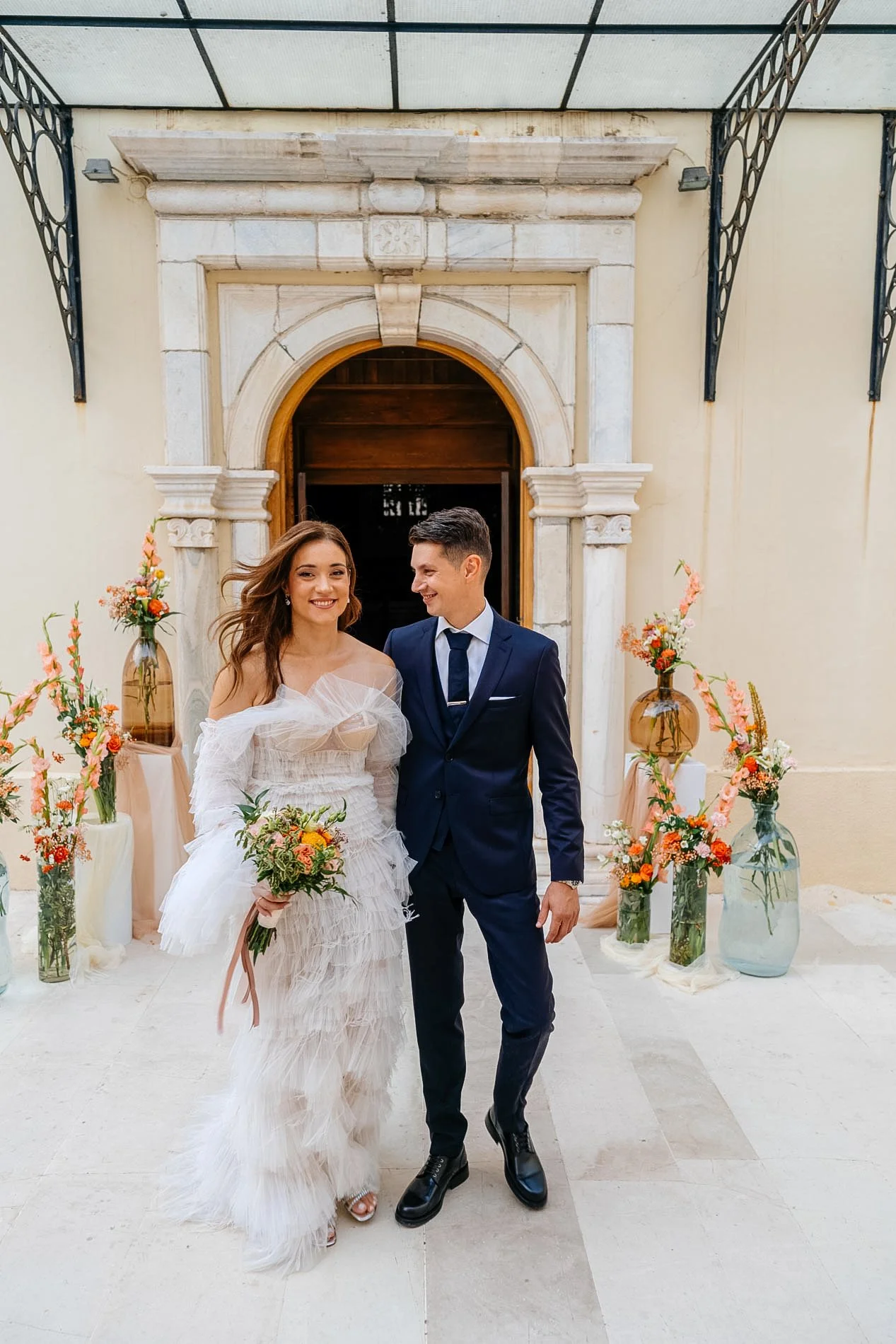 A newlywed couple smiling and walking hand-in-hand during their wedding ceremony, surrounded by floral arrangements in glass vases, in front of an ornate stone doorway.