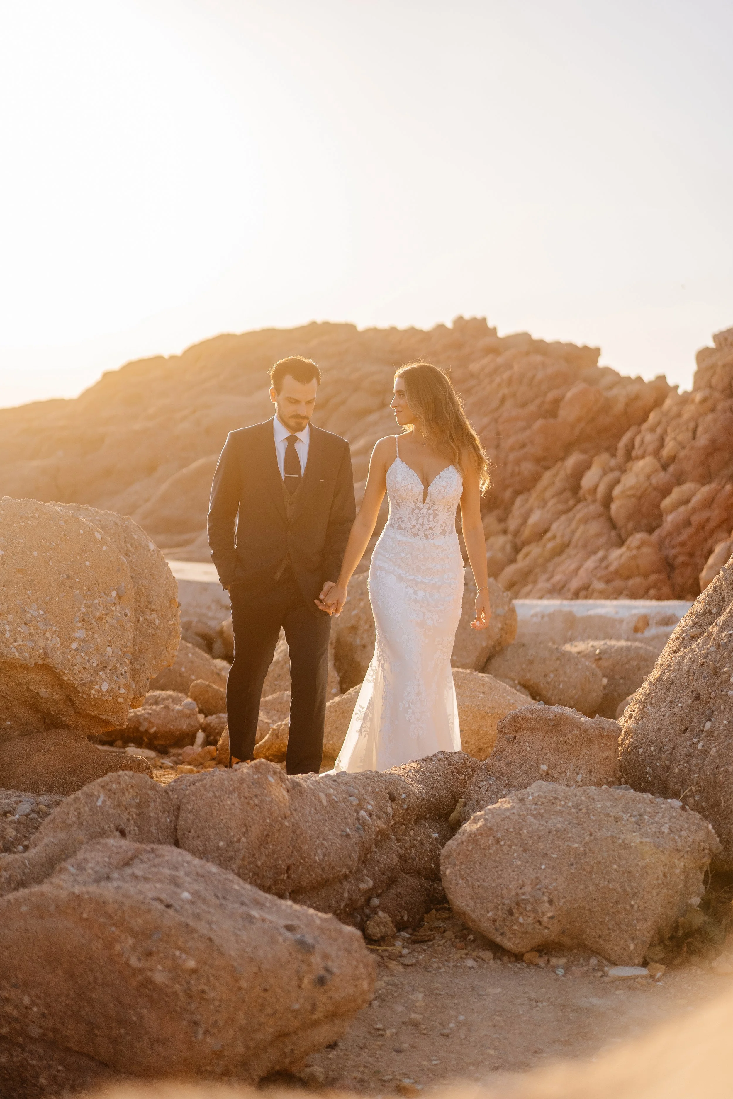 A bride and groom walking hand in hand among large rocks in a desert landscape during sunset.