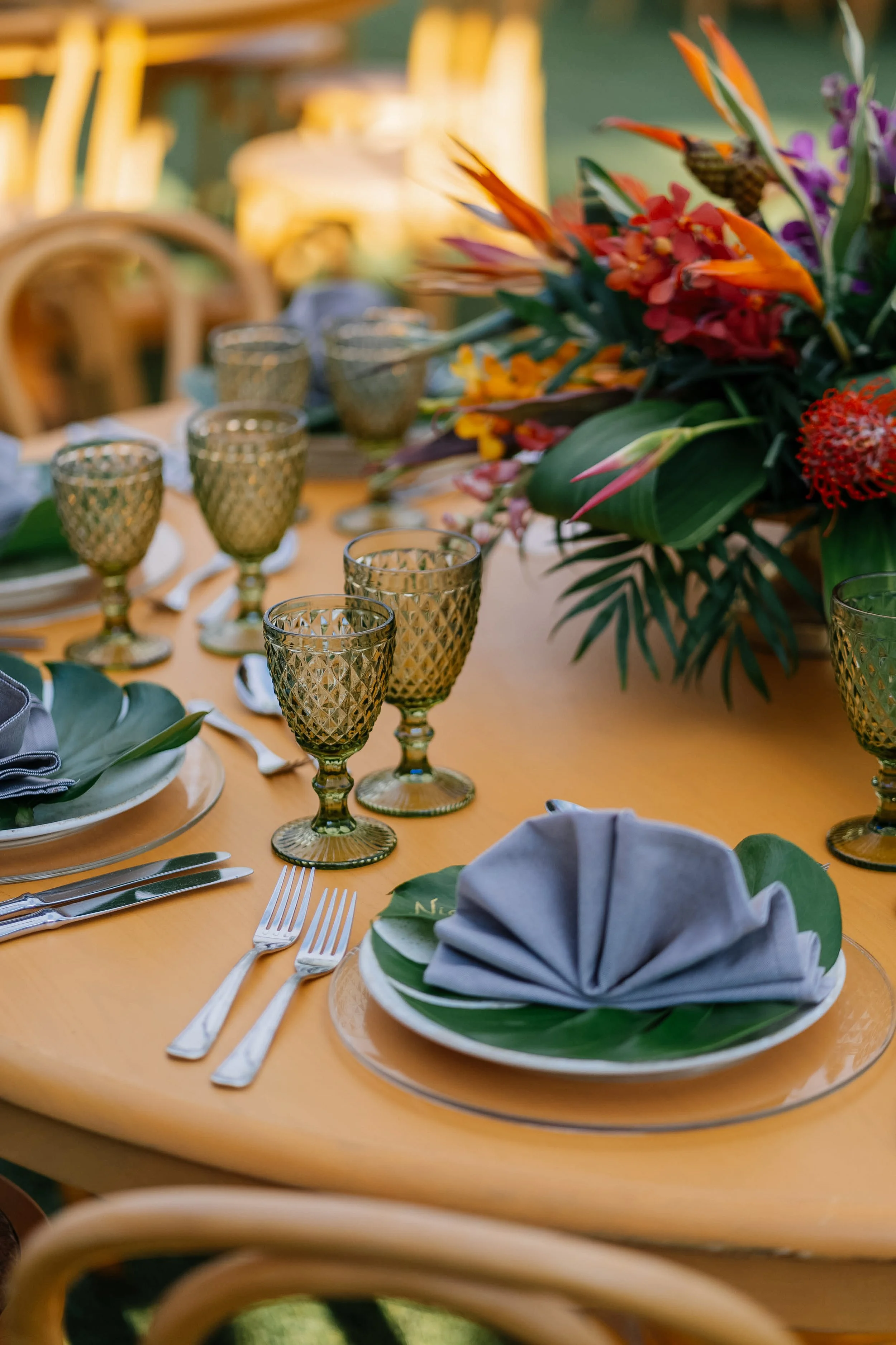 A table set for dining with glassware, plates, silverware, and cloth napkins arranged as a formal setting, with a large floral centerpiece featuring tropical flowers.