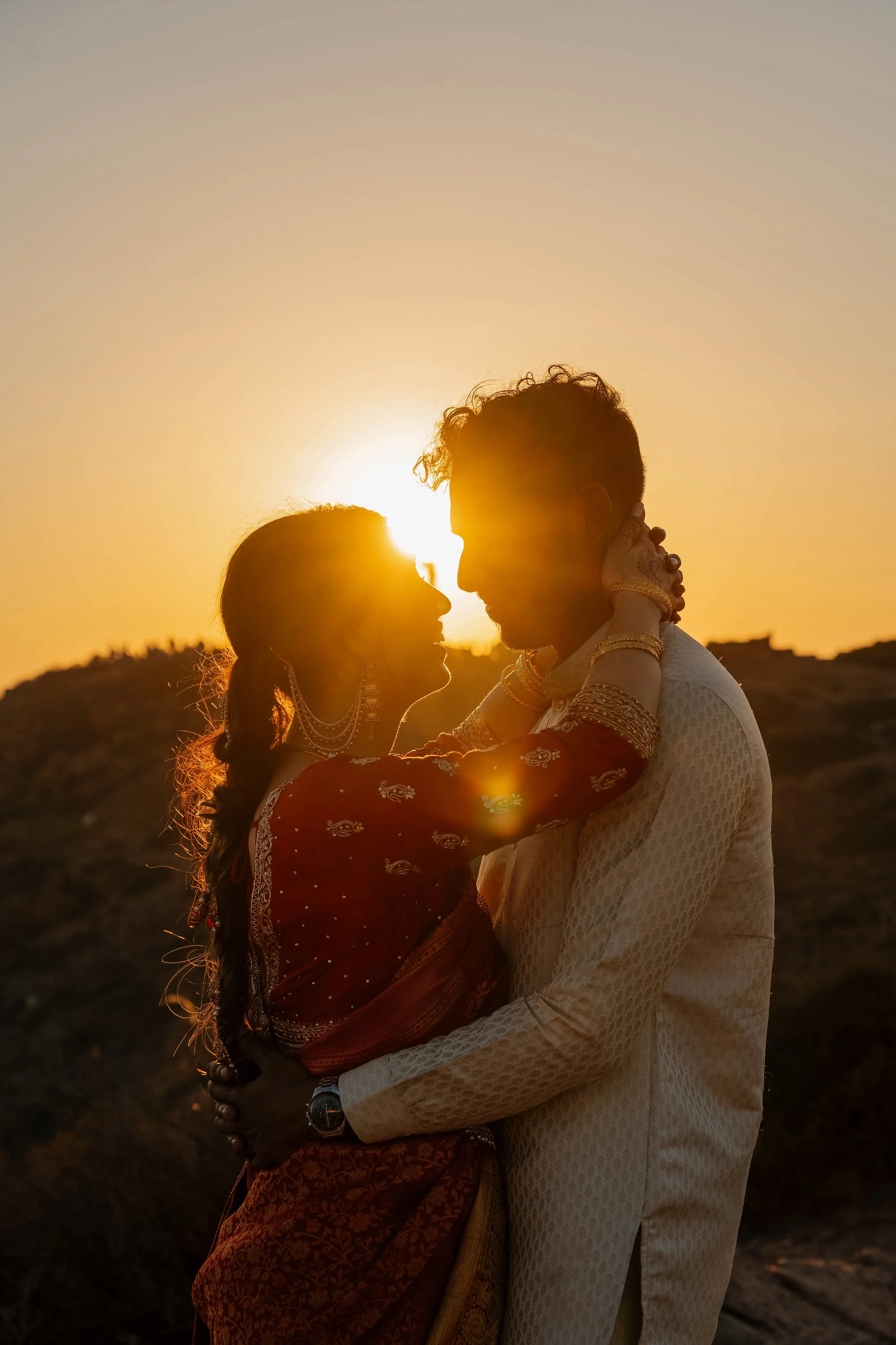 A couple dressed traditionally, holding each other and about to kiss during sunset, with a hilly landscape in the background.