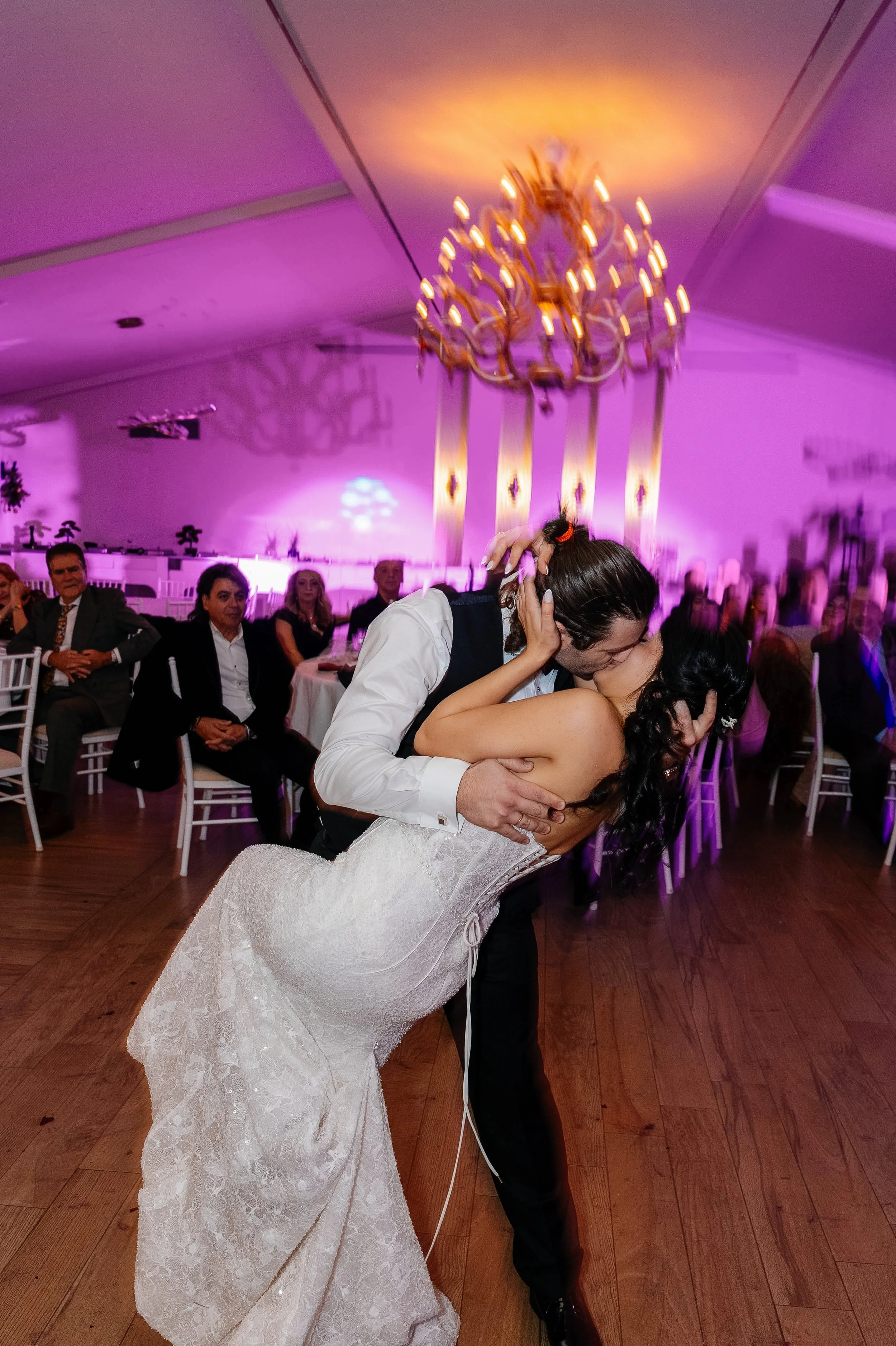 A bride and groom are dancing at a wedding reception, kissing passionately while the bride's dress flows on the wooden floor, with wedding guests seated in the background and a chandelier hanging from the ceiling.