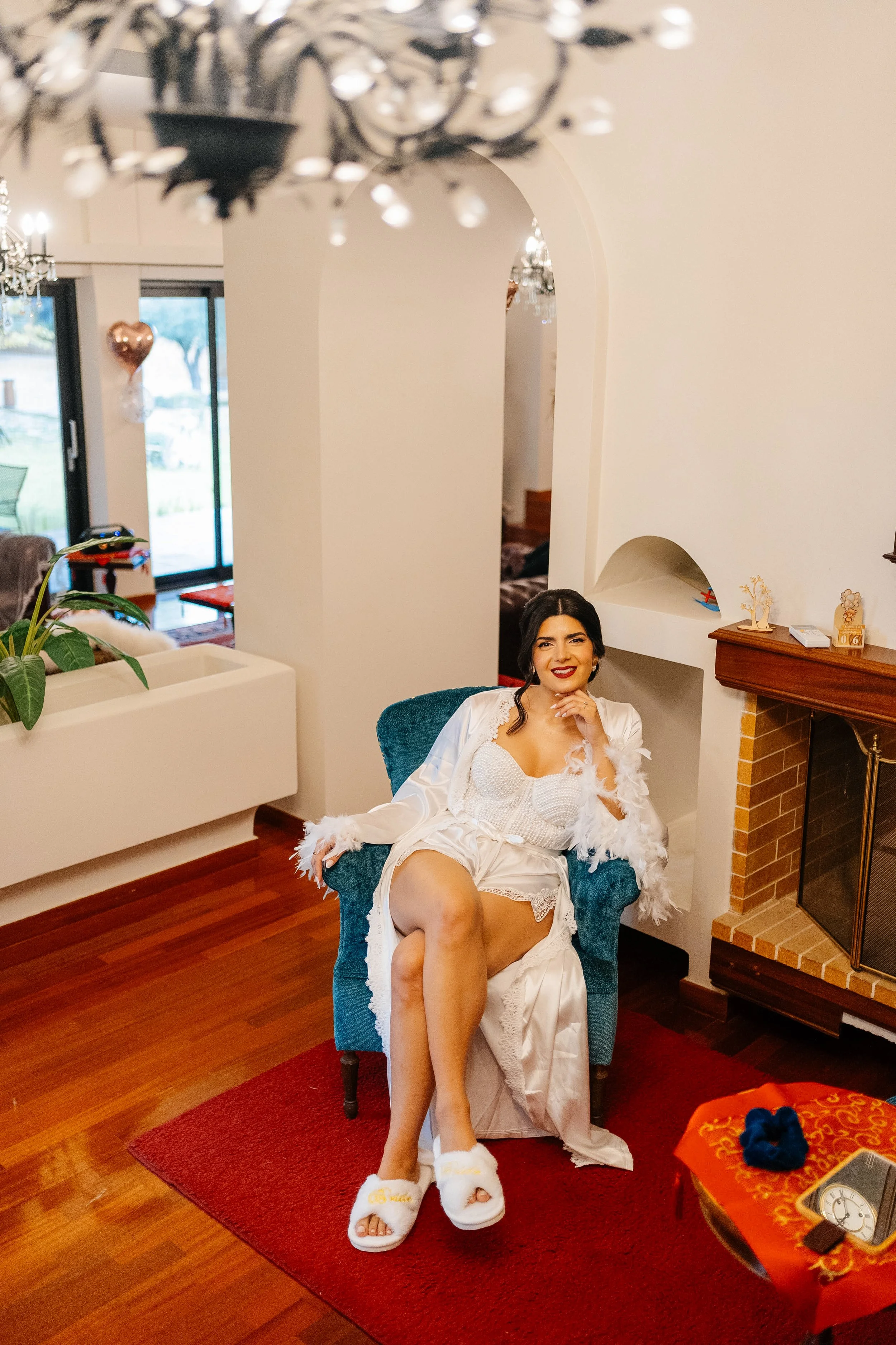 A woman sitting in a blue armchair, wearing a white satin robe with feather trim, slippers, and smiling at the camera with one leg crossed over the other, in a living room with wooden flooring, a fireplace, and festive decorations.