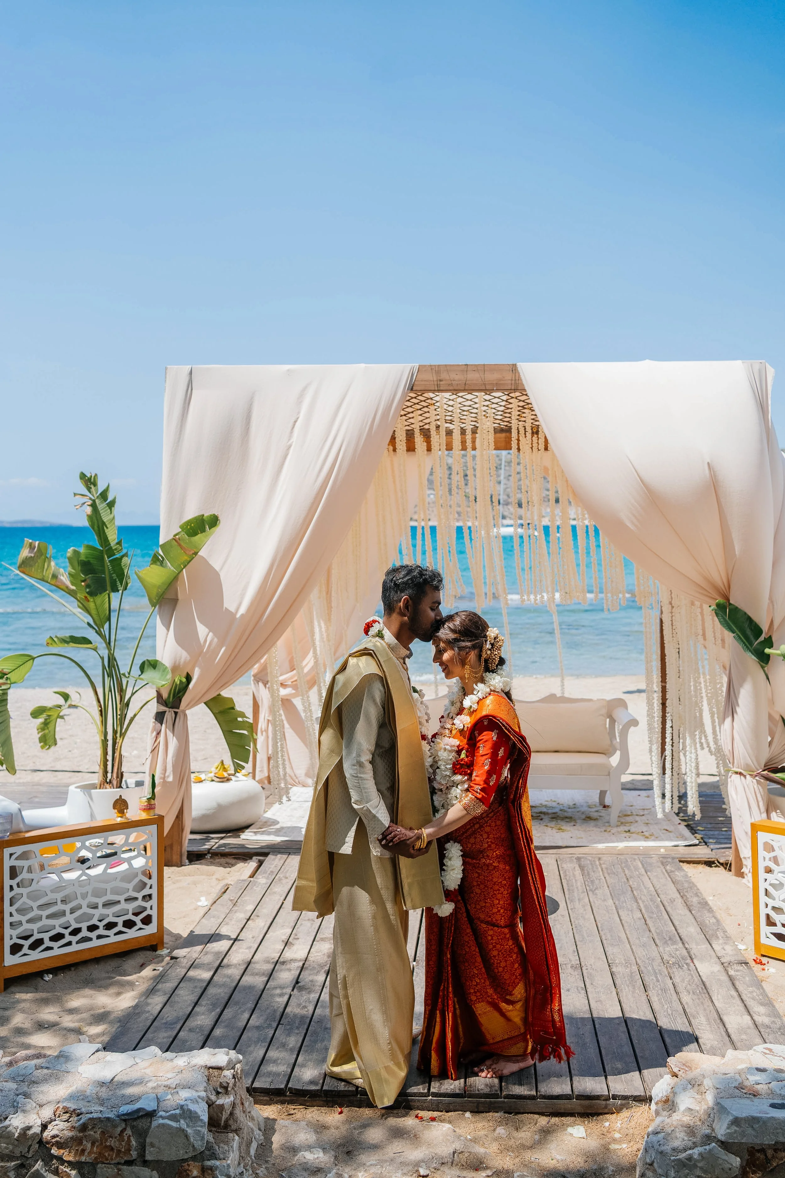 A couple wearing traditional Indian wedding attire kissing during a beach wedding ceremony under a beige canopy with hanging decorations, with ocean and blue sky in the background.