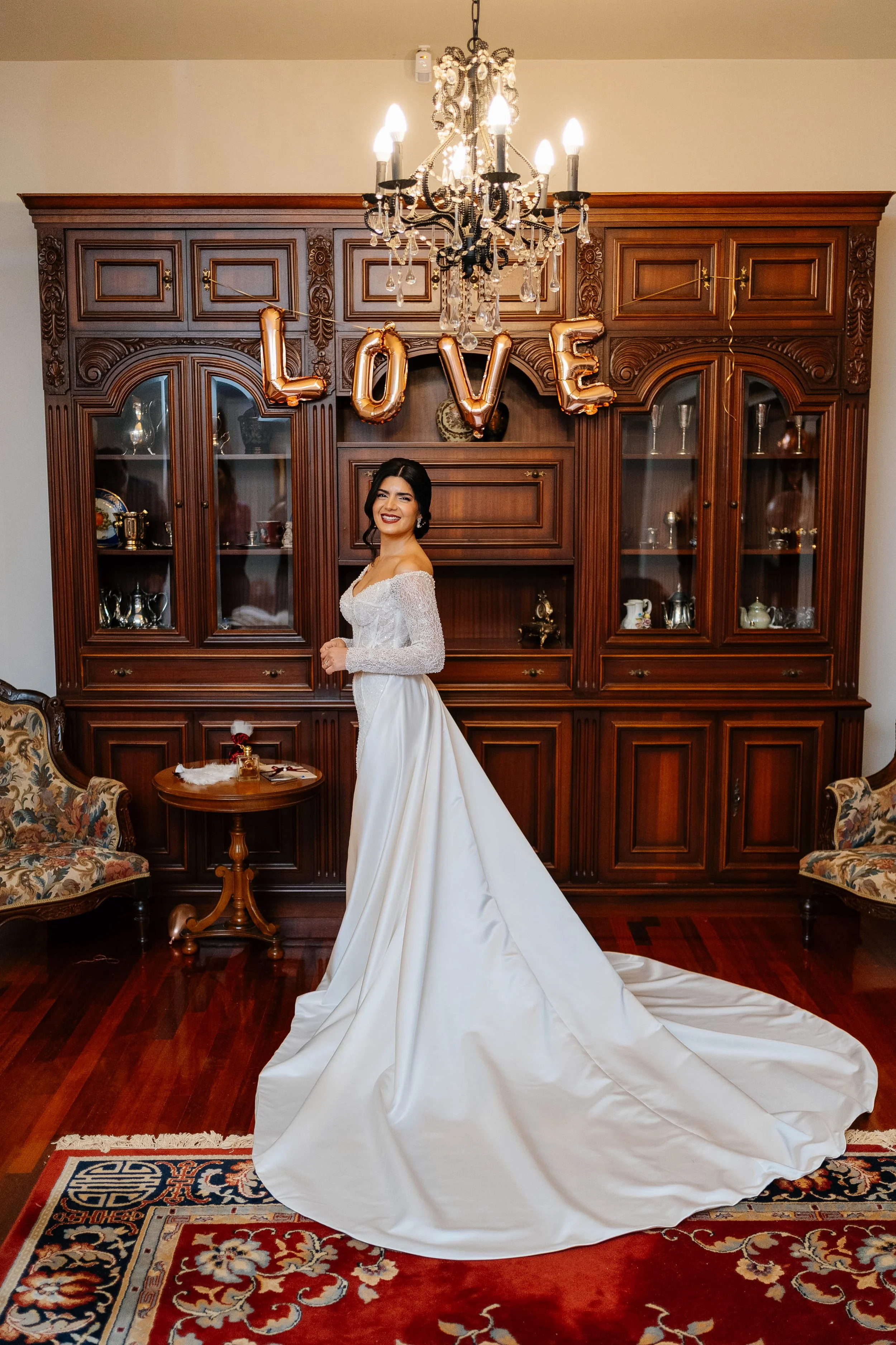 A woman in a wedding dress standing in a living room with a large wooden cabinet, a chandelier, and a red patterned rug. The word 'LOVE' in rose gold balloons is hanging from the chandelier.