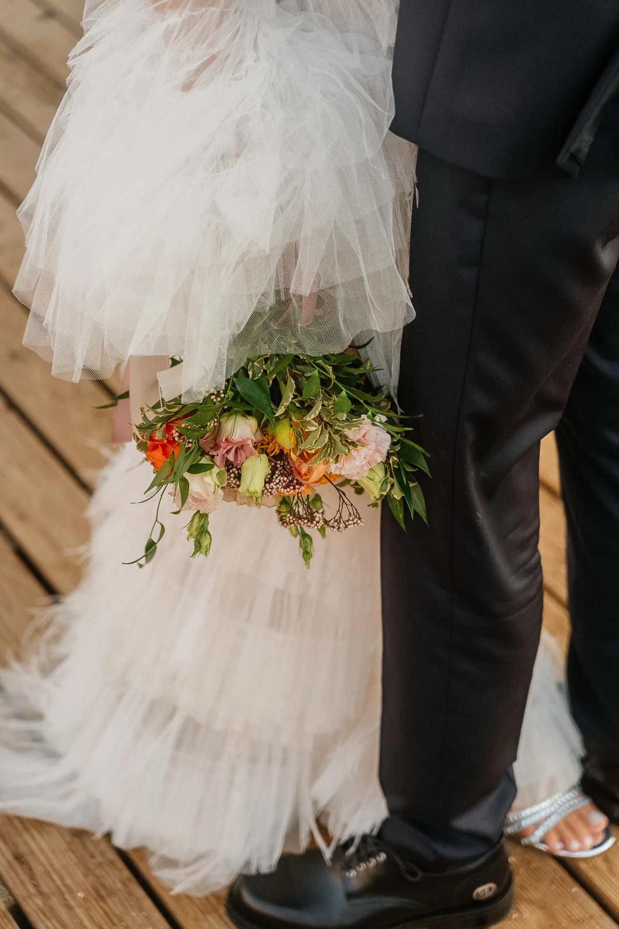 A bride wearing a white wedding dress with tulle layers, holding a colorful bouquet of flowers, standing next to a man dressed in black pants and shoes, on a wooden floor.