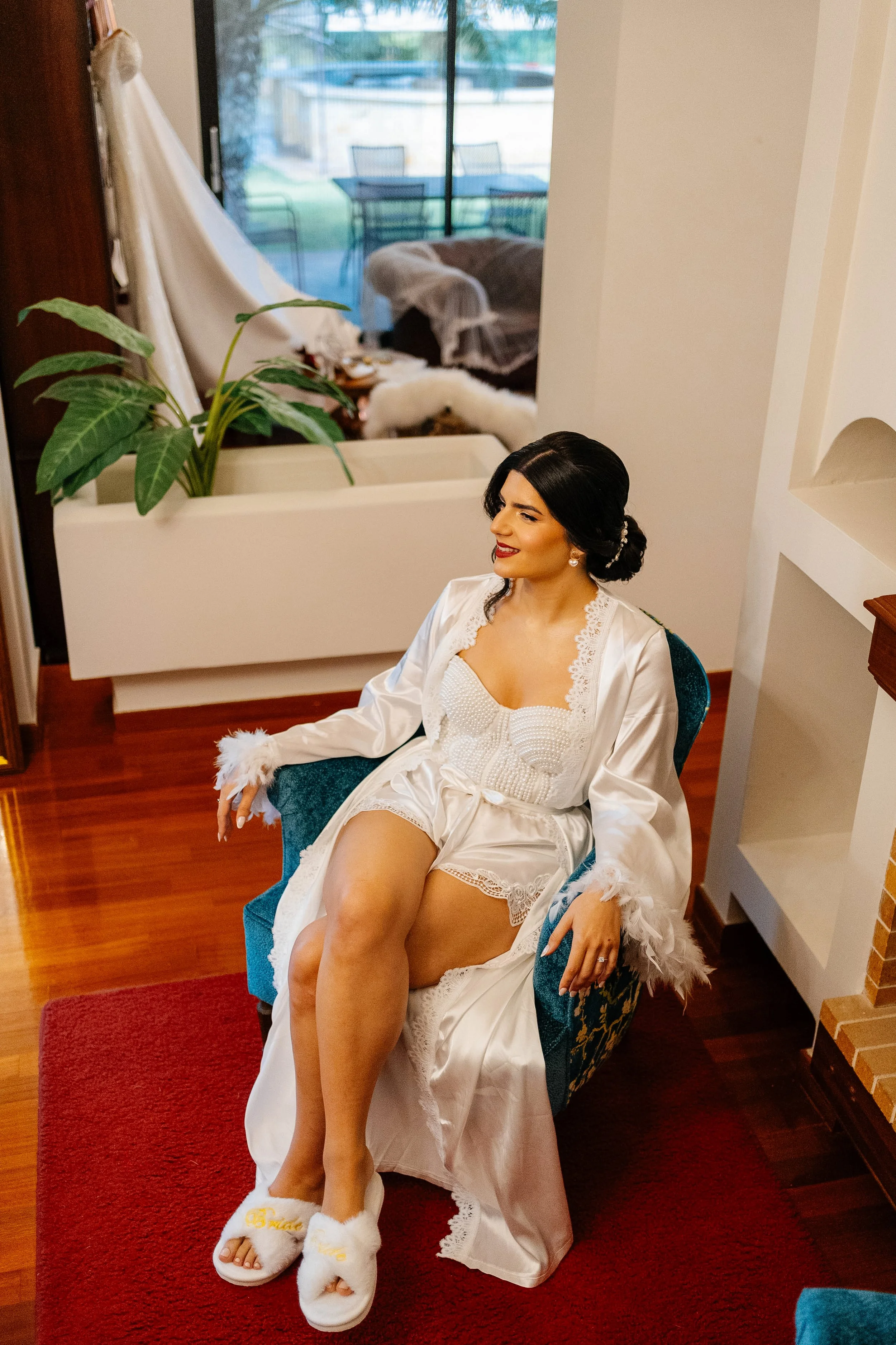 A woman dressed in white satin robe with lace trim, seated in a green chair, with legs crossed, smiling and looking to the side. She is wearing white fluffy slippers with gold embroidered words, and has dark hair styled in a low bun with a decorative