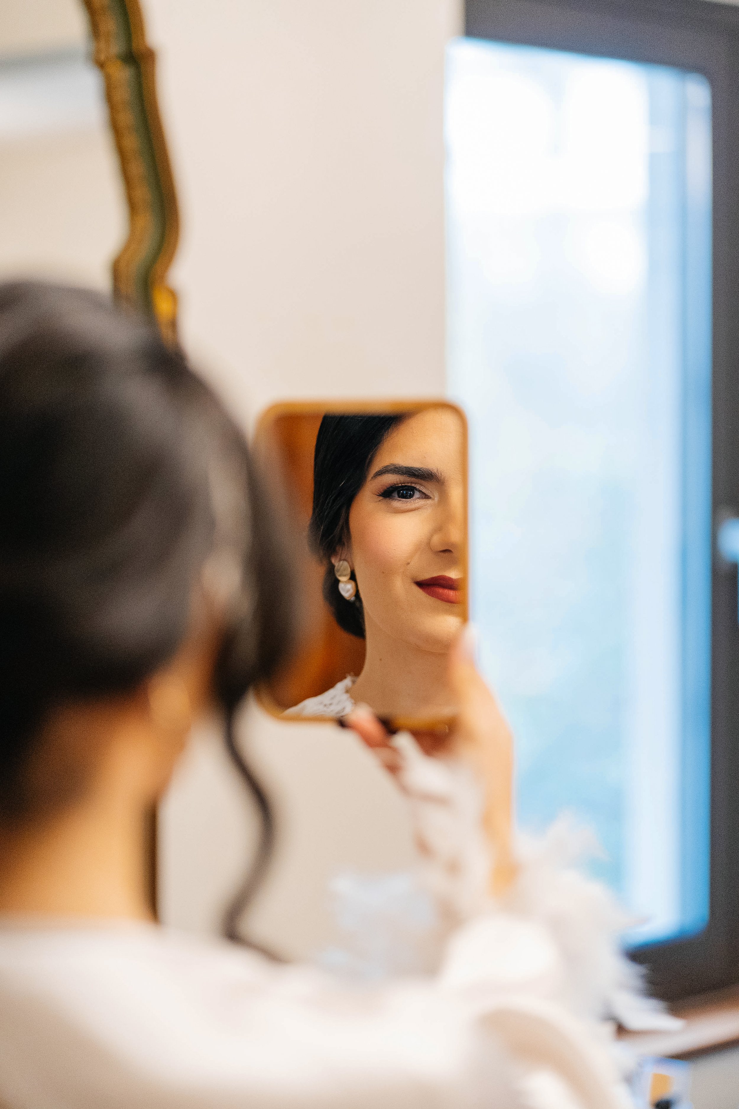 A woman looking into a small mirror with a partial view of her face reflected, wearing earrings and a white top, in a room with a bright window.